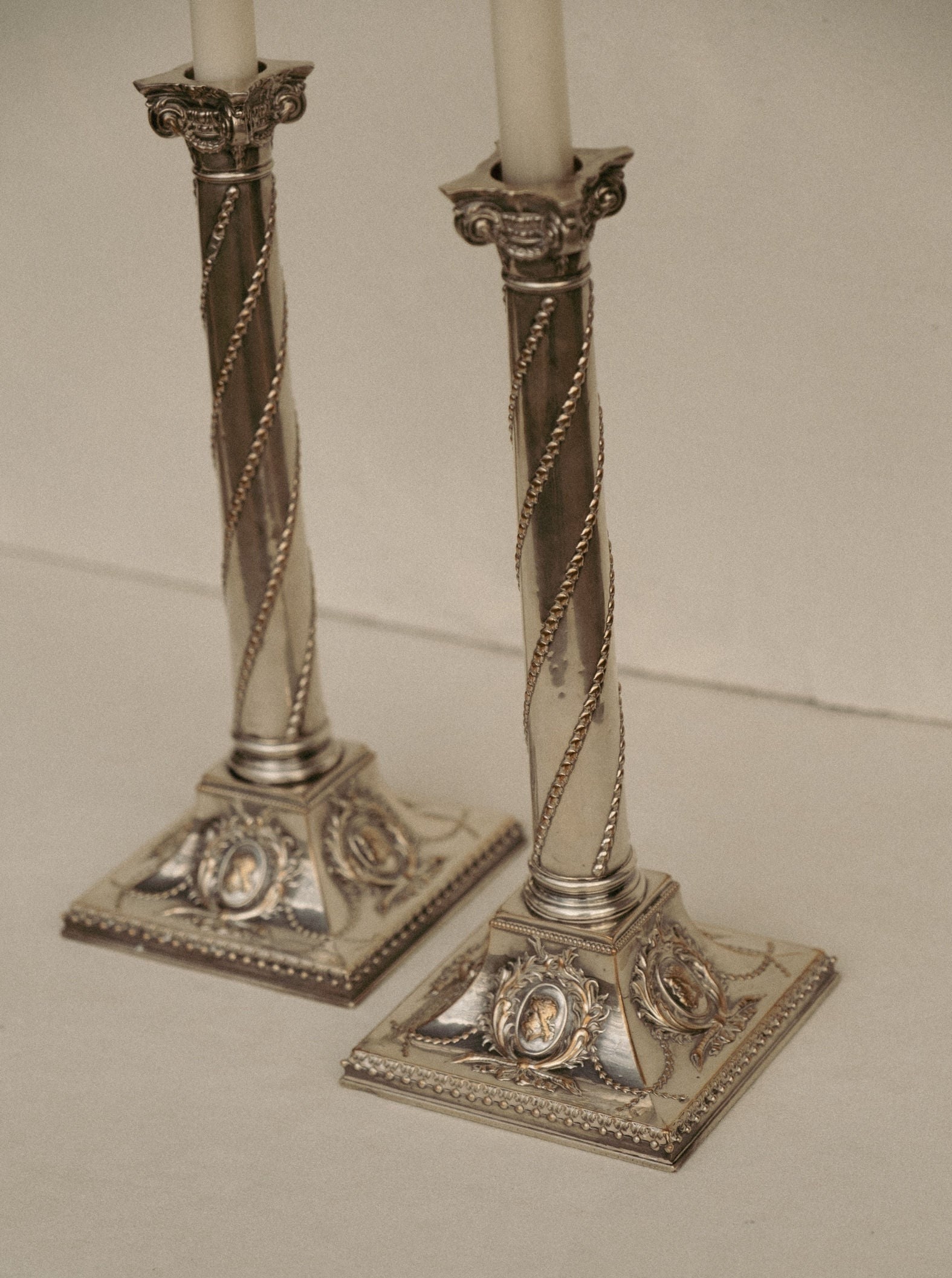 Two ornate, Casita Amada Pair of Column Sheffield silver plated candle stick holders with a twisted design and decorative details on the base are placed side by side on a plain, beige surface. The antique candle stick holders have unlit, white candles. The intricate patterns and craftsmanship highlight a classical, elegant style.