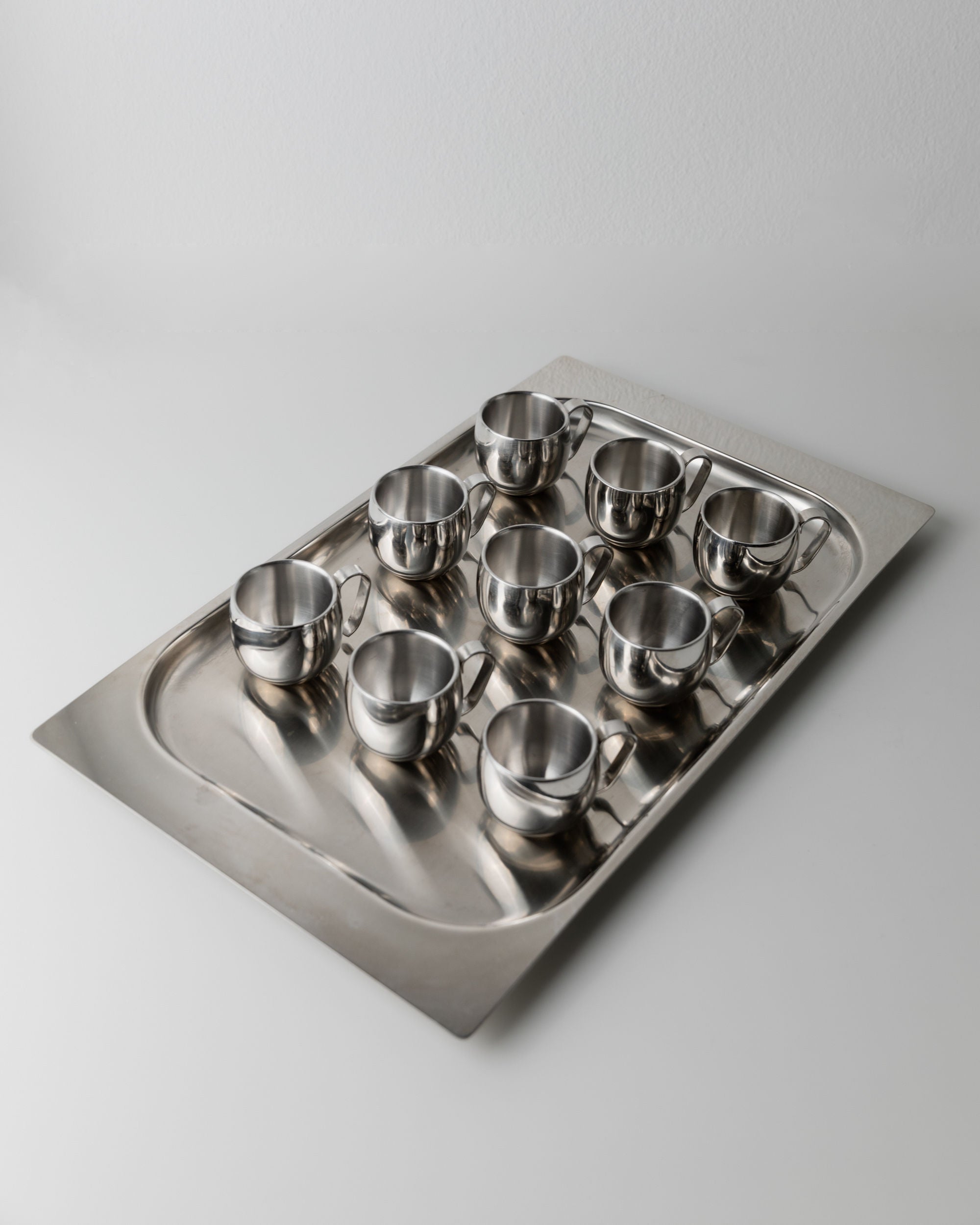 The Coffee set by IMCO 70s from Bottega Jacobs features a stainless steel tray holding ten sleek, shiny metal cups on a smooth white surface. Its minimalist design enhances the aesthetic appeal and vintage charm.