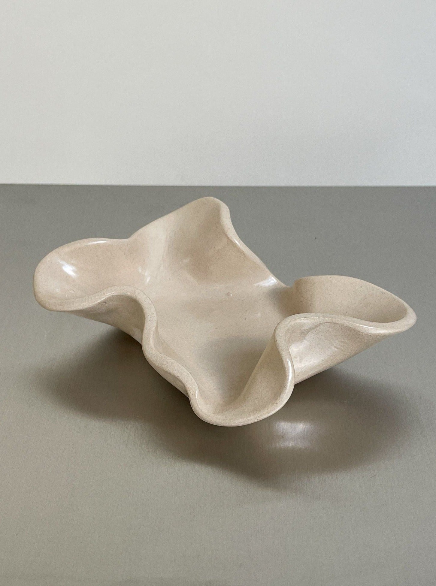 The Soapholder in stoneware by 2222Studio features a cream, wavy ceramic design with an irregular shape—perfect as a stylish, handmade touch for your bathroom decor. Displayed on a smooth gray surface against a light background.