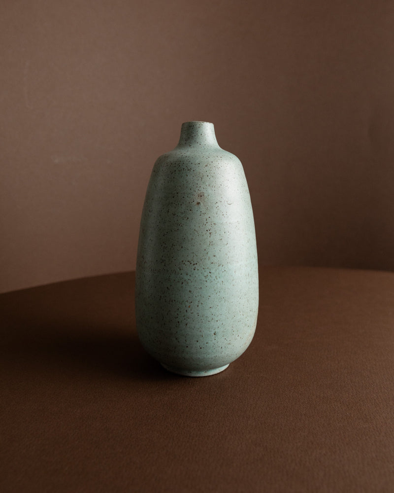 The Mint-Colored Vase by Carl Fischer (1960s) for Bottega Jacobs features a simple, rounded vintage design with a narrow neck and speckled light blue-green glaze, shown on a brown surface against a brown background.