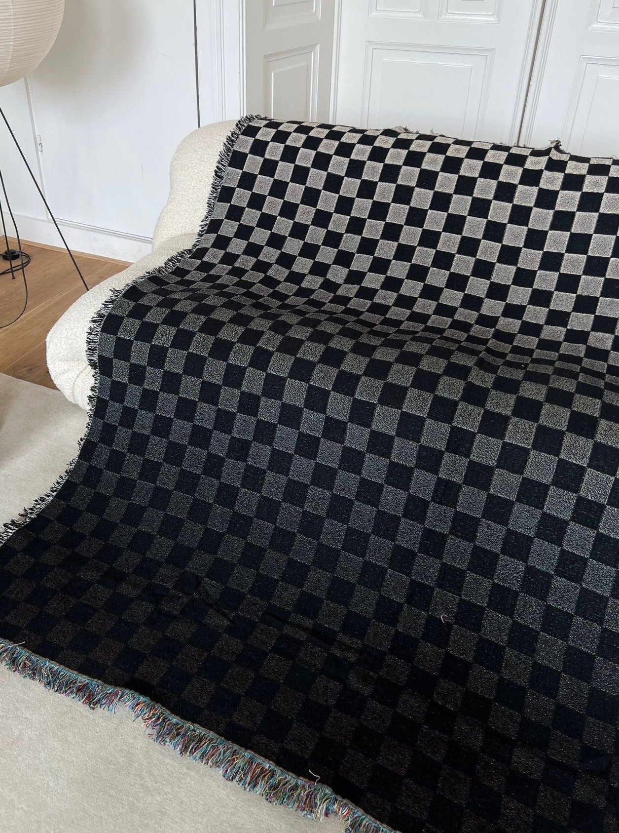 A cream sofa is adorned with the 2222Studio Iki Cotton Jacquard Throw, featuring black and gray patterns and fringed edges. The versatile throw covers the seat and backrest. White panel doors and a standing lamp are seen in the background.