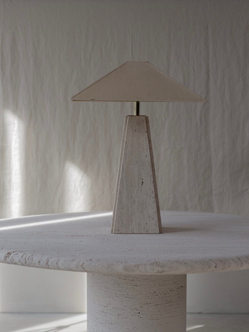 The Bicci de' Medici Camille Breesch - Travertine Table Lamp graces a round textured white table with its minimalist design, featuring a tapered travertine and brass base, matching triangular shade, and casting soft shadows on the light-colored wall.