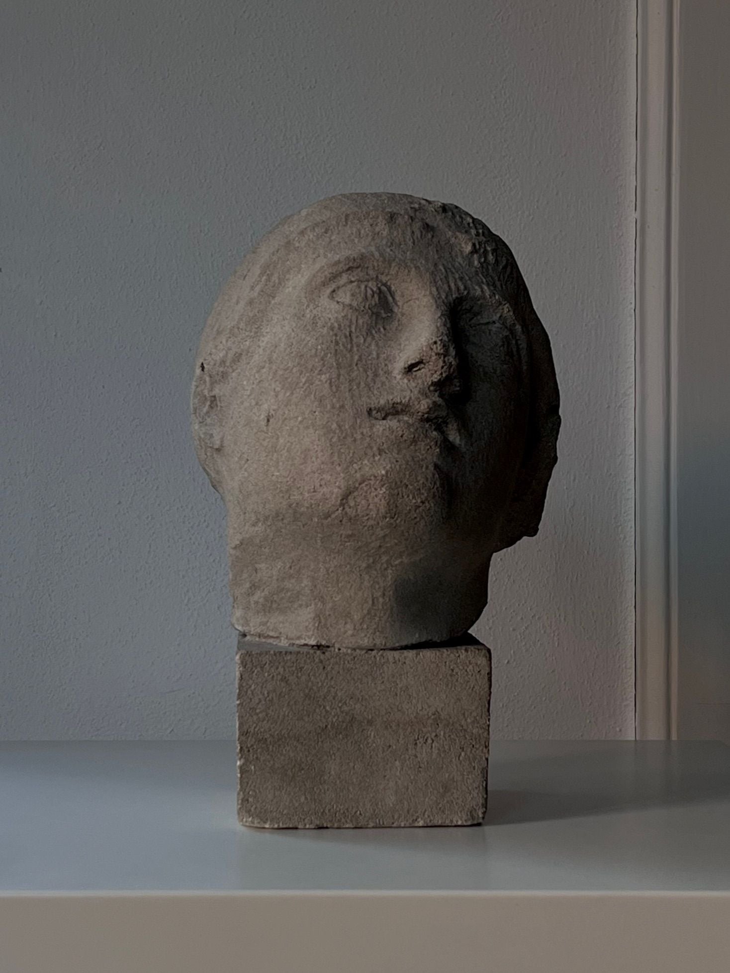 A Decorative Bust by Médecine, crafted from gray sandstone and featuring a human head design, is set on a square base. Placed against a simple wall, it radiates mid-20th-century charm, with soft shadows accentuating its textured patina.