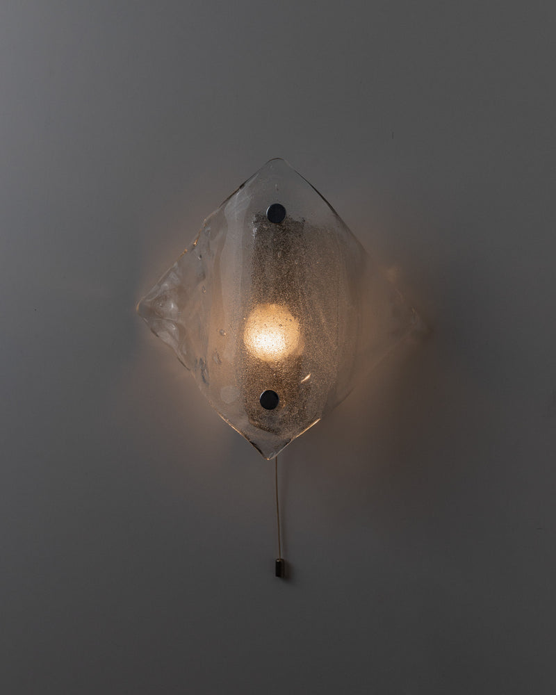 The Vintage Murano Glass Wall Lamp by Kalmar from Bottega Jacobs features a diamond-shaped, textured glass cover that emits soft, warm light. Mounted on a gray wall, it includes a hanging cord switch and embodies the elegance of space age design.