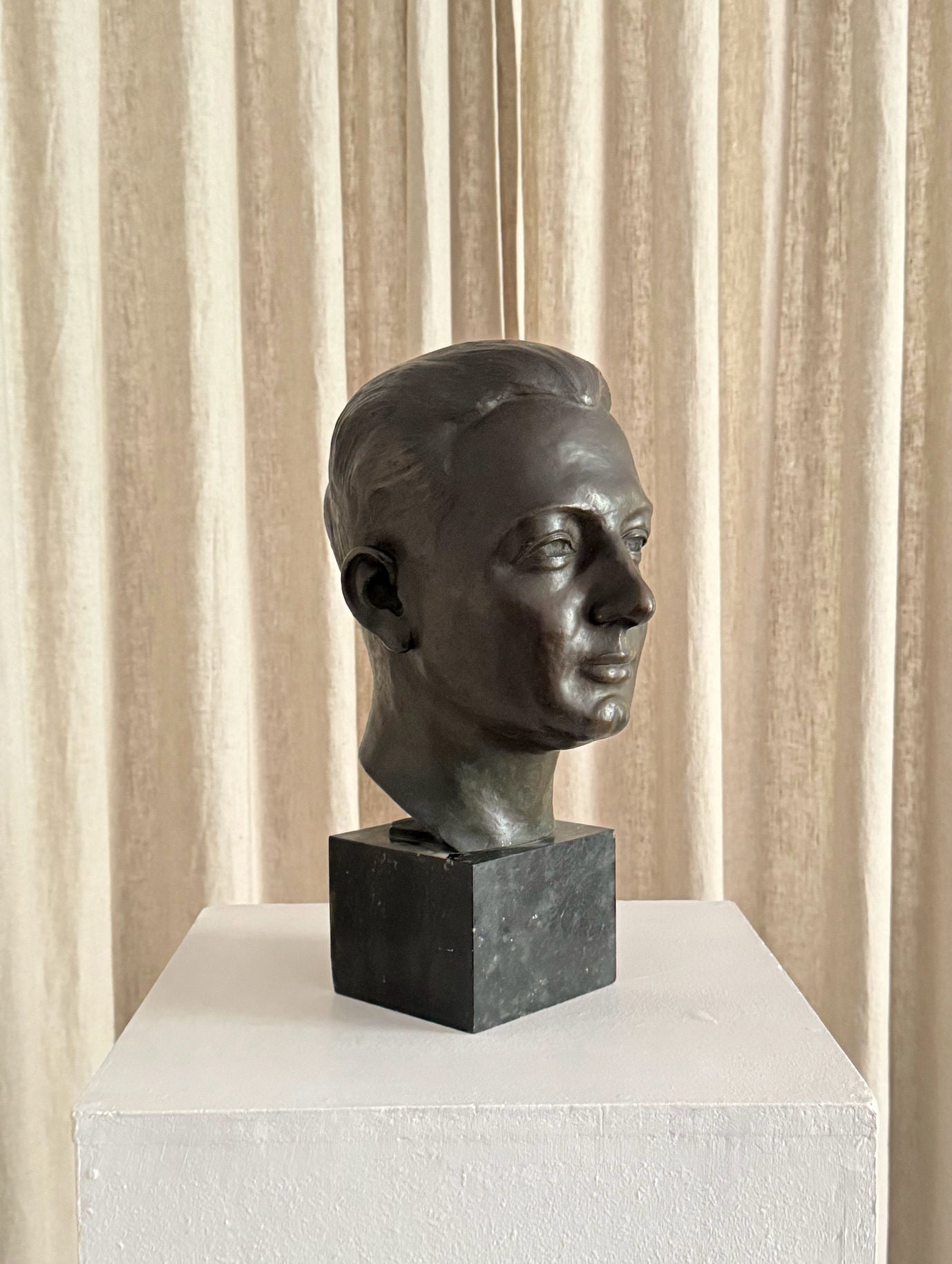 The "Testa Virile" Sculpture 1930s by FEBRERO SHOP is a bronze bust of a short-haired man gazing right, displayed on a dark pedestal and white plinth, set before elegant beige curtains.
