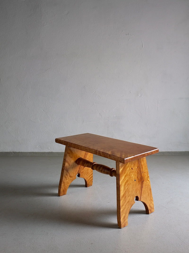 A small wooden stool carved from solid Karelian birch wood with a polished finish featuring a simple design, a flat rectangular seat, and two curved legs connected by a horizontal spindle. The Rustic Solid Karelian Birch Stool | Latvia | 1950s by Veter Vintage is placed on a smooth, light gray floor against a plain, light-colored wall.