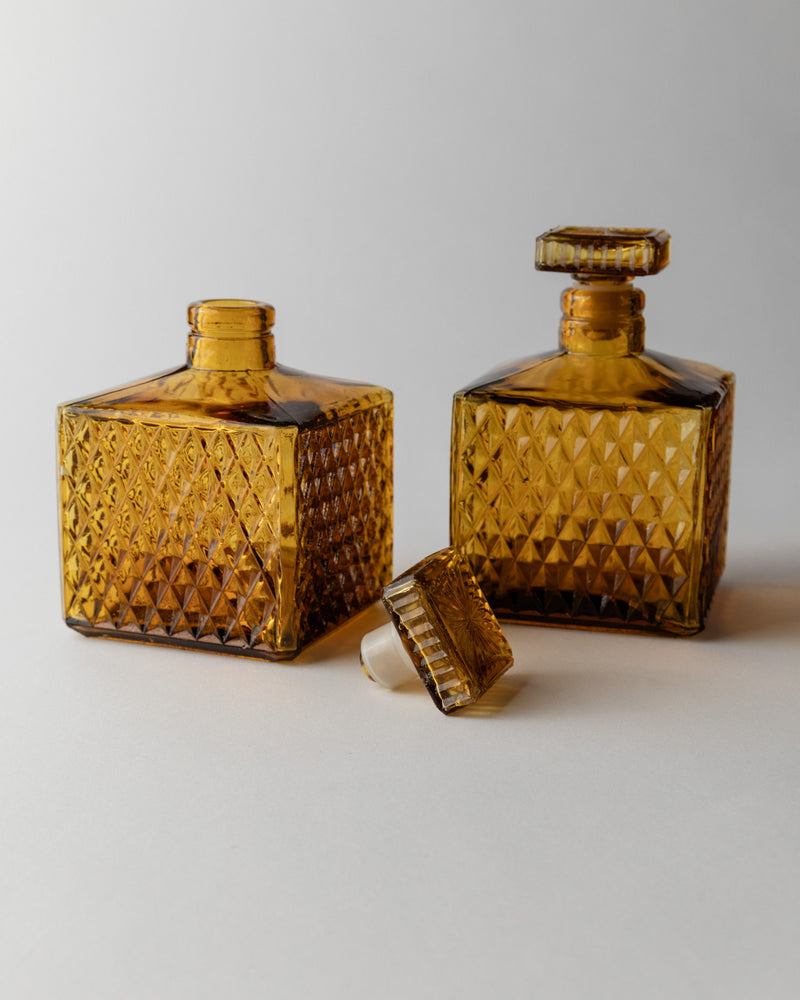 Two Bottega Jacobs Amber Glass Decanter 70s pieces feature square, textured patterns in vintage style; one has a matching stopper, while the other's stopper rests in front, all set against a neutral background.