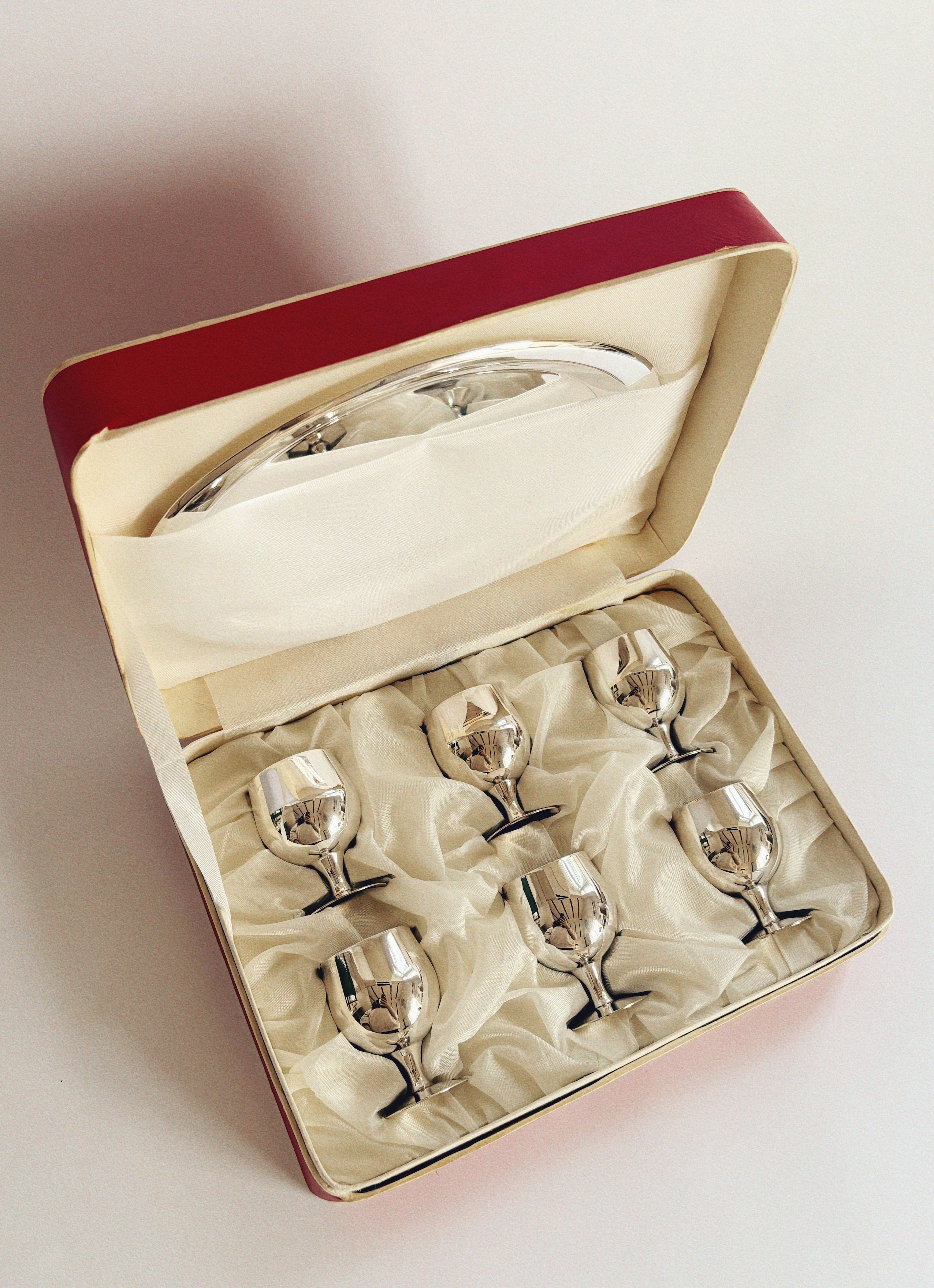 The Huerto "Silver Plated 70's Liquor Server," an exquisite set of six silver-plated liquor glasses, is showcased in a cream-colored satin-lined red box that evokes collectible items from communist-era Poland, complete with a matching silver tray gracefully positioned against the lid.