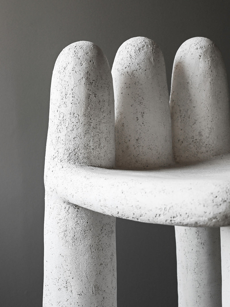 The Washed Concrete Chair by Studio OSKLO features a sculptural, finger-shaped backrest and a textured, stone-like surface—unique handmade furniture for indoor or outdoor use, shown against a plain gray background.