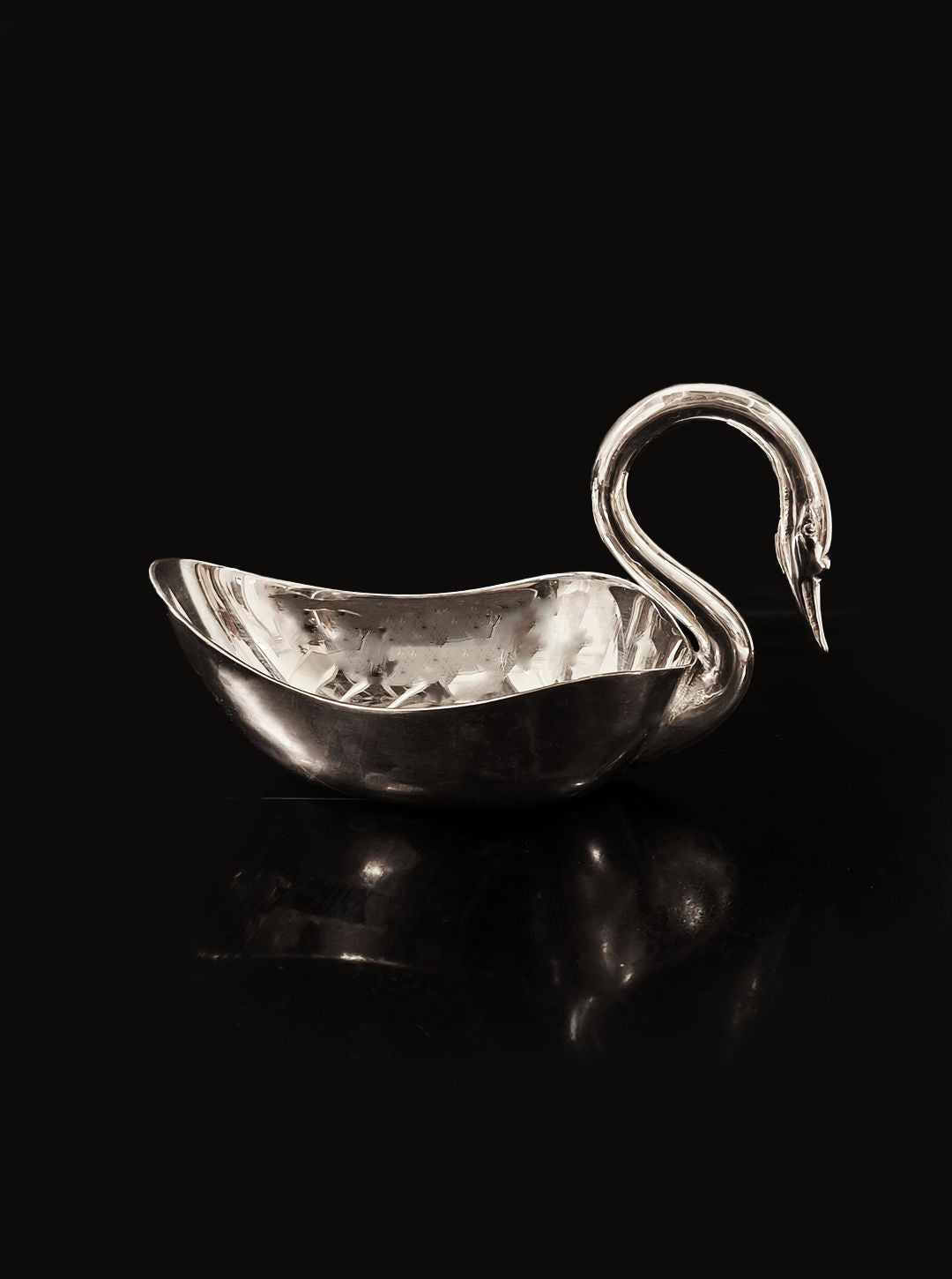 Introducing the Swan Silver Saucer by Les Objoies, a stunning decorative serving piece. This silver bowl is elegantly designed in the shape of a swan, featuring a graceful, curved neck and a smooth, reflective surface that beautifully contrasts against its black background. The saucer's textured interior enhances its sleek exterior elegance.