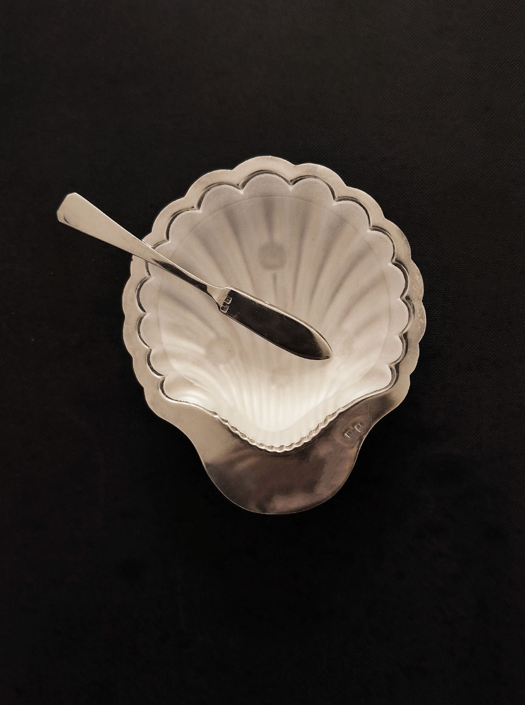 A Les Objoies Shell Butter Dish, scallop-shaped and silver-plated with a matching spreader knife, gleams elegantly on a dark surface.