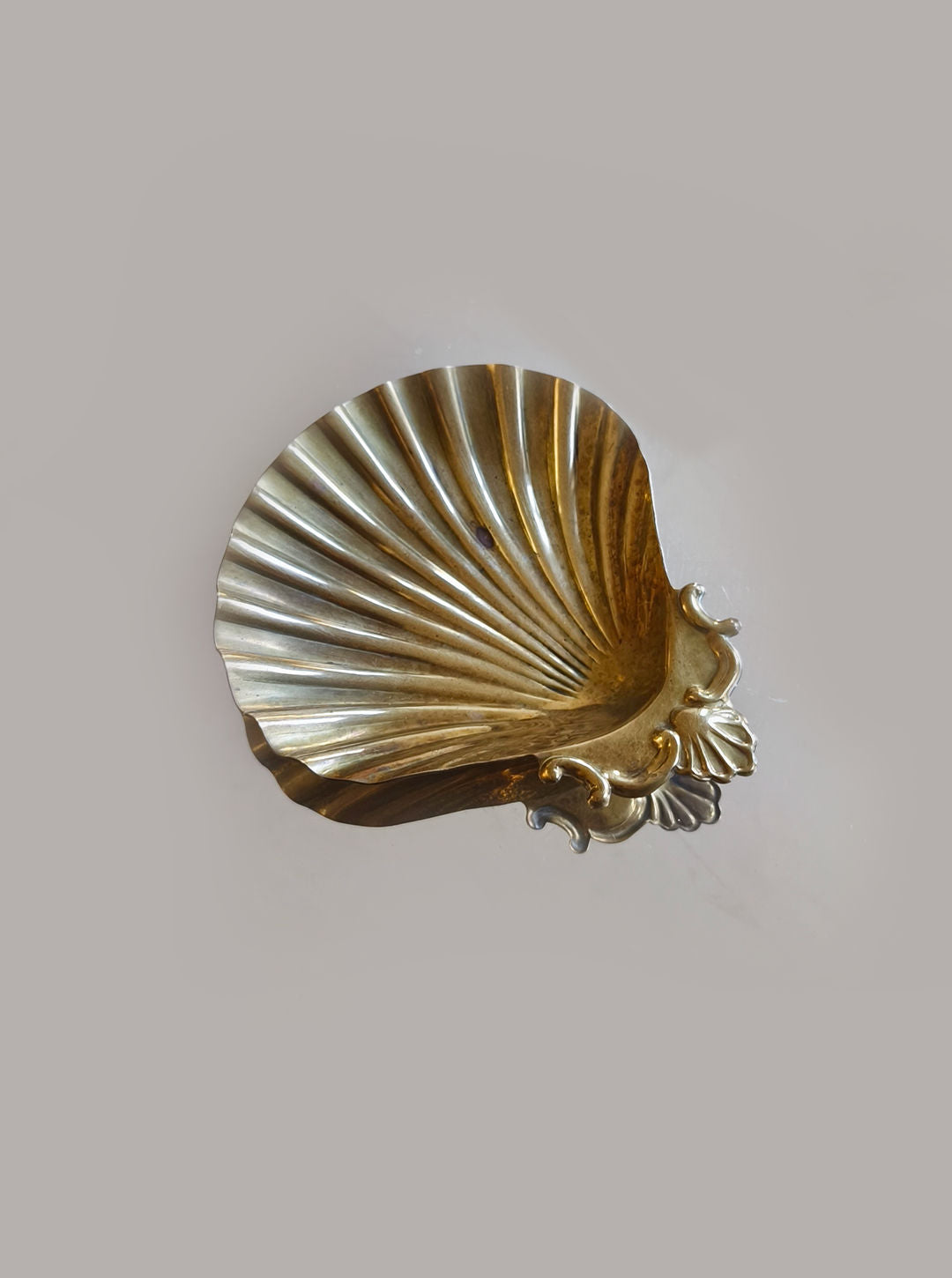 The Shell Brass Vide Poche by Les Objoies is a decorative brass seashell-shaped piece with intricate leaf details on the bottom edge and a golden hue, set against a plain gray background.