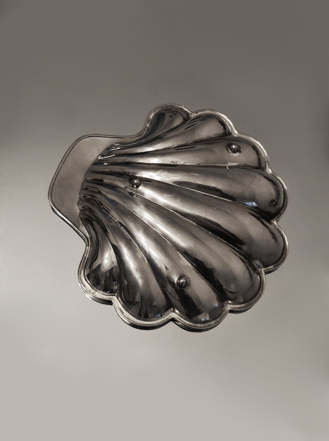 The Shell Shape Vide-poches by Les Objoies is a silver-plated metal dish resembling a seashell with a reflective surface, featuring a scalloped edge and smooth finish, elegantly presented on a neutral background.