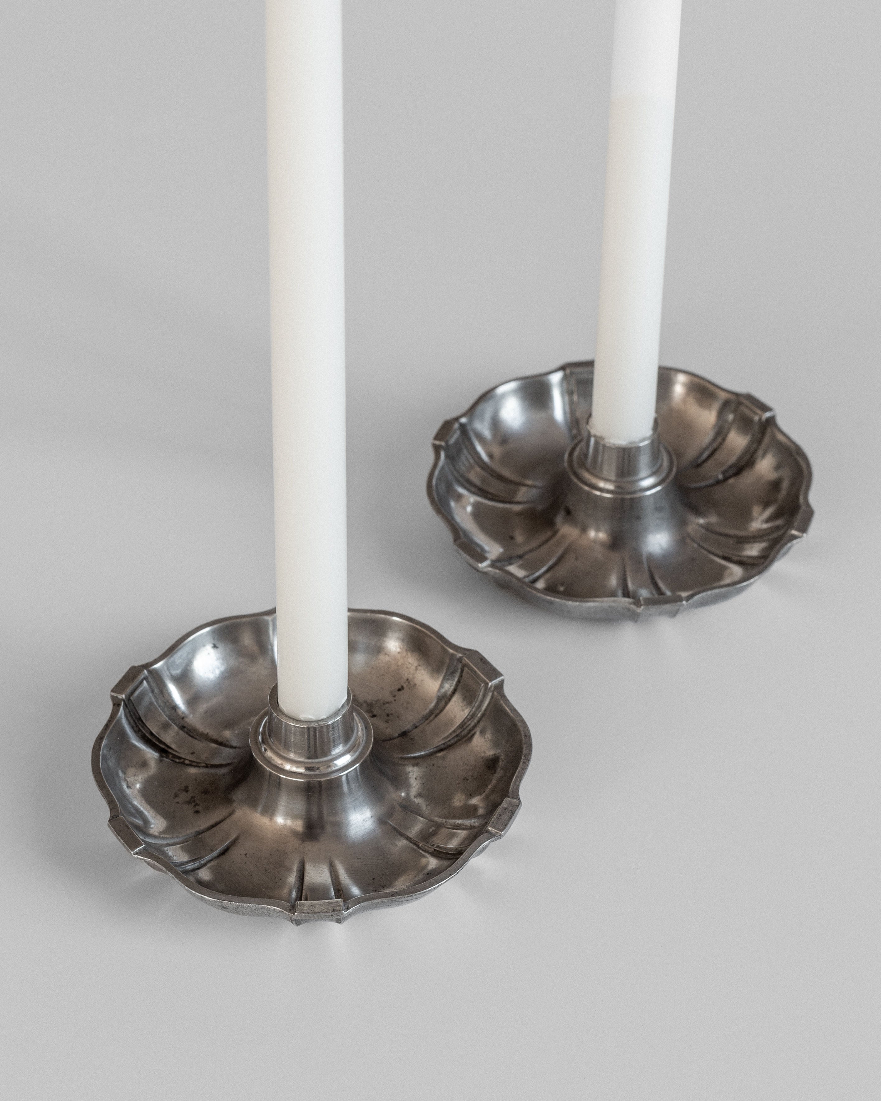 Two white candles rest in the elegant Spigel Modernist Metal Candleholders from the 1930s, their scalloped silver design reminiscent of C.G. Hallberg's Swedish Modernist metalwork gracing a light surface. The candleholders' shiny finish and vintage design lend an air of sophistication to the setting.