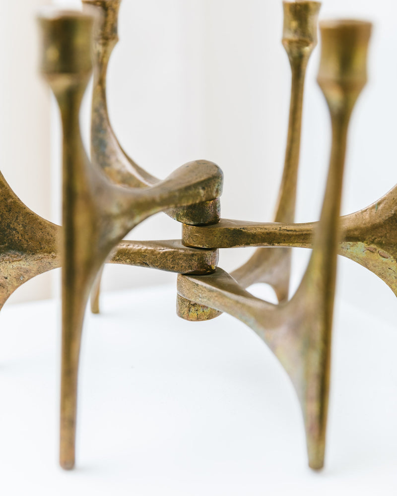 A close-up of the Candleholder by Michael Harjes in Bronze 60s, offered by Bottega Jacobs, features a vintage three-legged design with interlocking metal arms that highlight its modern and abstract geometry. The aged patina stands out beautifully against a softly blurred background.