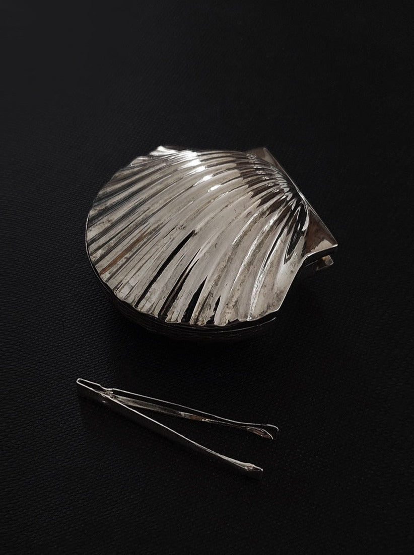 A silver, textured Small Shell Box jewellery holder with a clamshell design lying open on a dark surface, accompanied by a pair of silver tweezers positioned below it. Brand: Les Objoies.