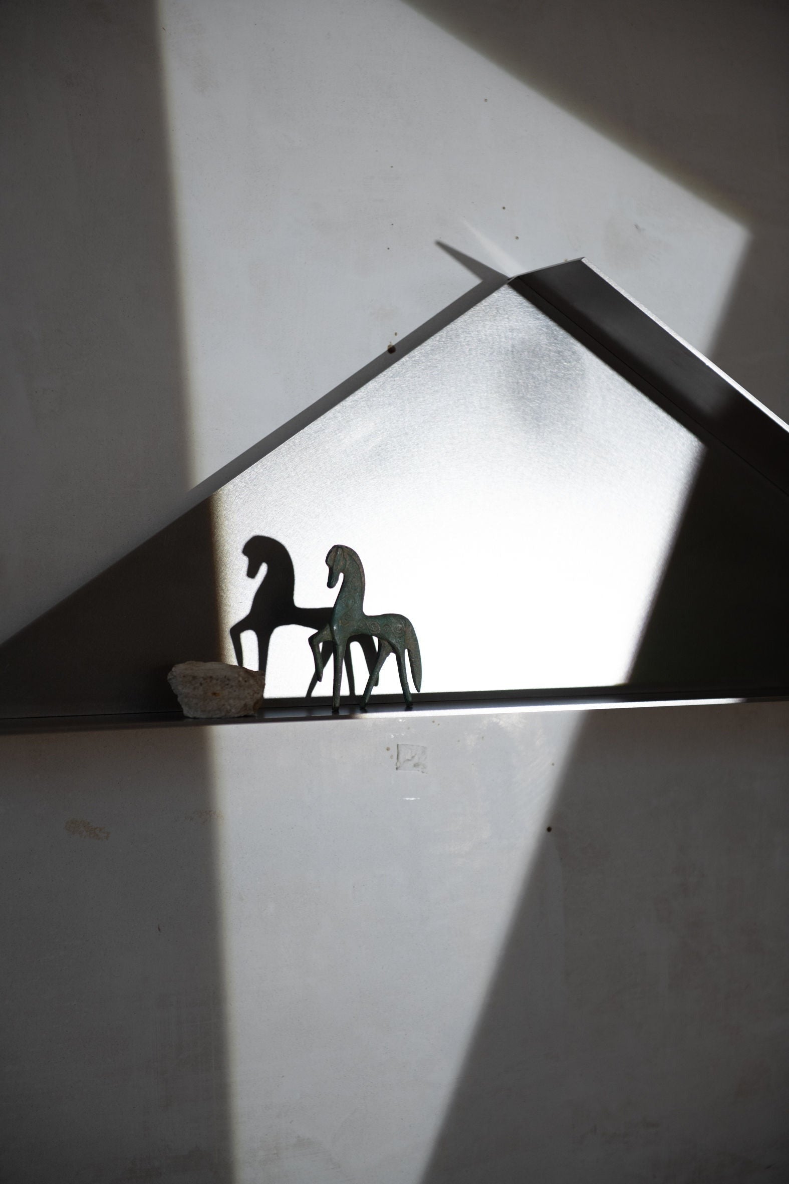 A minimalistic décor display featuring two sculptural horse figures on a COSEINCORSO The Front - Shelf. The scene is illuminated by angular beams of light, casting sharp shadows and highlighting the sleek, modern design. These limited edition pieces in stainless steel are set against a backdrop of neutral tones, adding to the contemporary aesthetic.