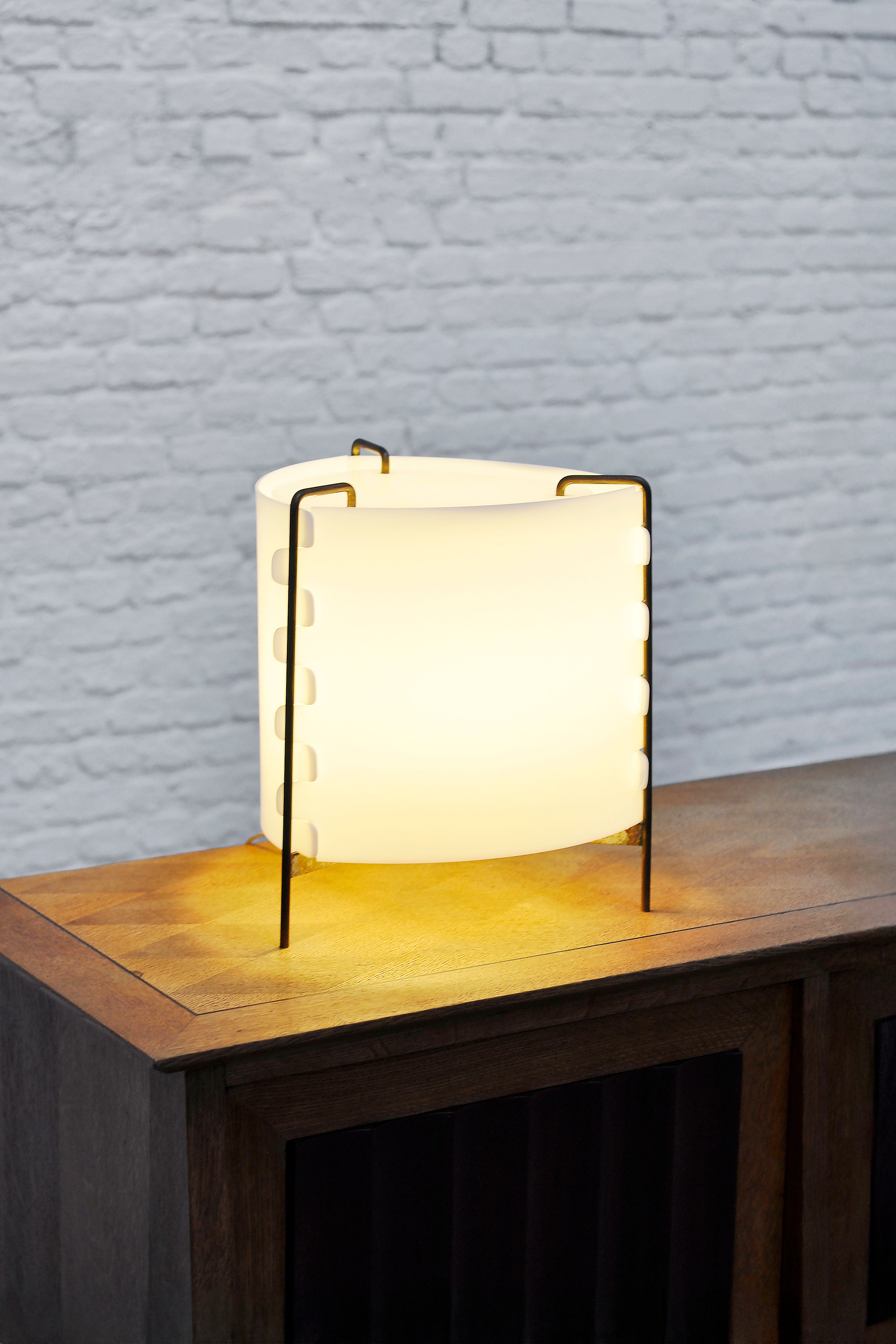 The M8 Fanal lamp by Joseph-André Motte for Huchers-Minvielle, sold under the Introverso brand, rests on a wooden table. Its cylindrical design with a white Perspex shade and metal frame emits a warm glow, perfectly enhancing the white brick wall and capturing French mid-century modern style.