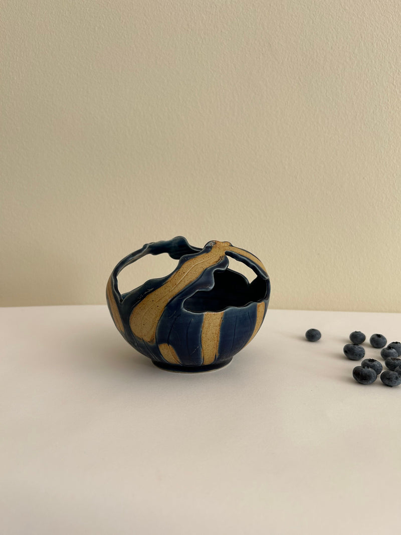 An Artisan Ceramic Bowl by Flock Studio, featuring a navy and tan stripy pattern with intricate wavy edges reminiscent of Balinese artistry, sits on a light surface. A few blueberries are scattered beside it, set against a plain beige wall.