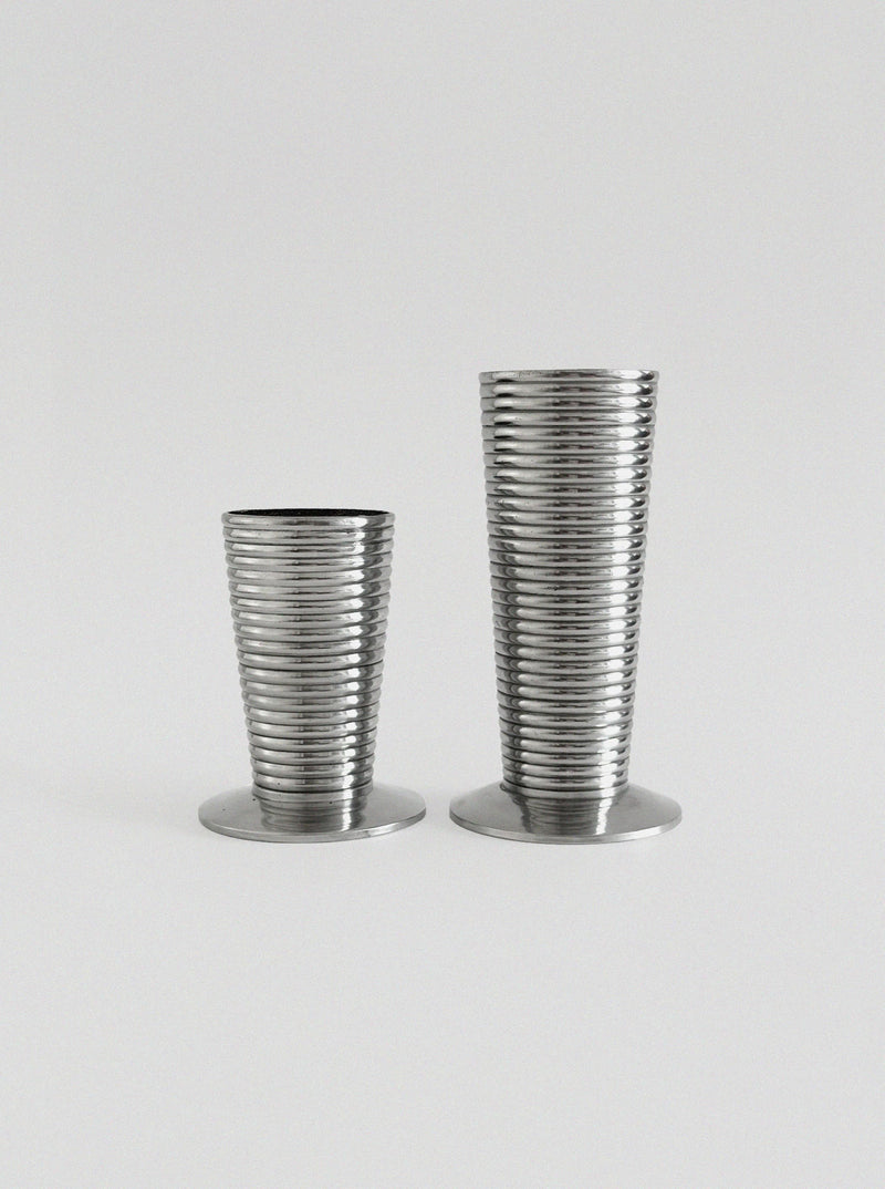 The Pair of Aluminum Vases by Vintage Stories features two ribbed silver vases of varying heights, displayed side by side on a light background—a sophisticated modern decor accent. The shorter vase is on the left, the taller one on the right.