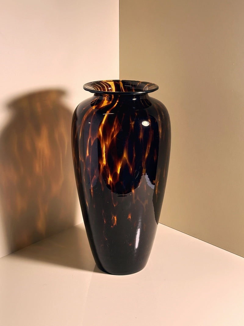 A glossy Tall Murano Tortoise Vase by Dodo Vintage casts a shadow on a light surface, where the neutral background and soft lighting highlight its exquisite tortoise speckle technique and showcase the vase's rich color and texture variations.