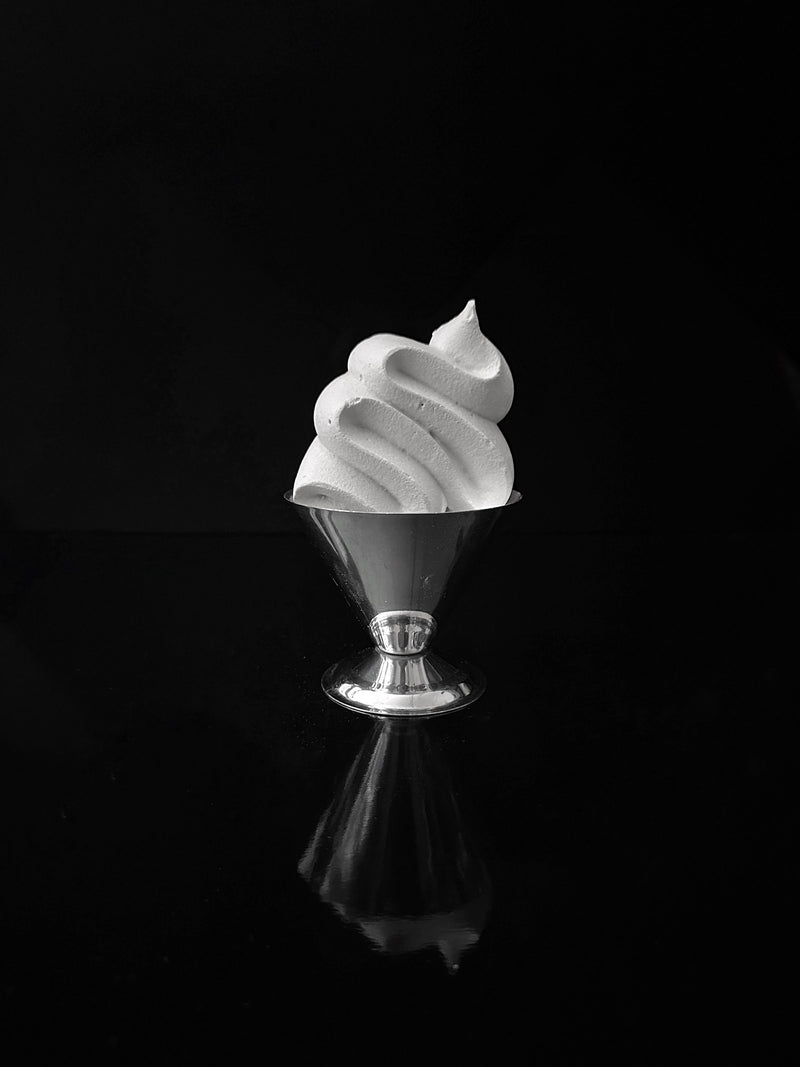 A grayscale image showcases a swirl of soft-serve ice cream in one of the "Set of 4 Italian Stainless Steel Dessert Cups" by Dodo Vintage, evoking memories of 1980s Italian dessert cups, set against a dark background. The glossy surface reflects both the ice cream and the cup beneath.