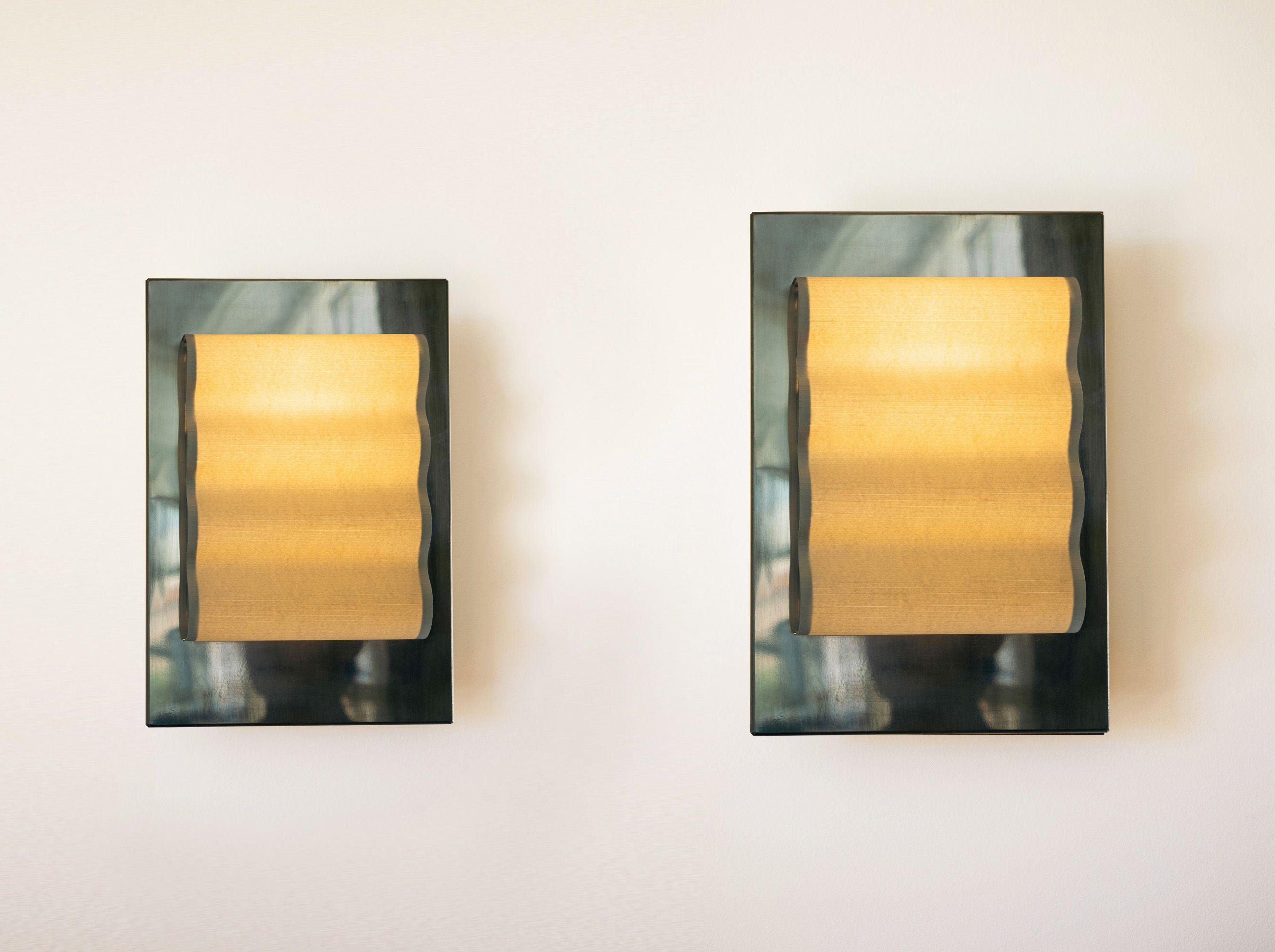 Introducing the Wall Light Frame Miroir M by Violaine d'Harcourt: a pair of handcrafted modern wall sconces featuring rectangular black frames and warm golden fabric shades. Mounted on a light-colored wall, these exquisite sconces emit a soft, ambient light that enhances minimalist design while creating a welcoming atmosphere.