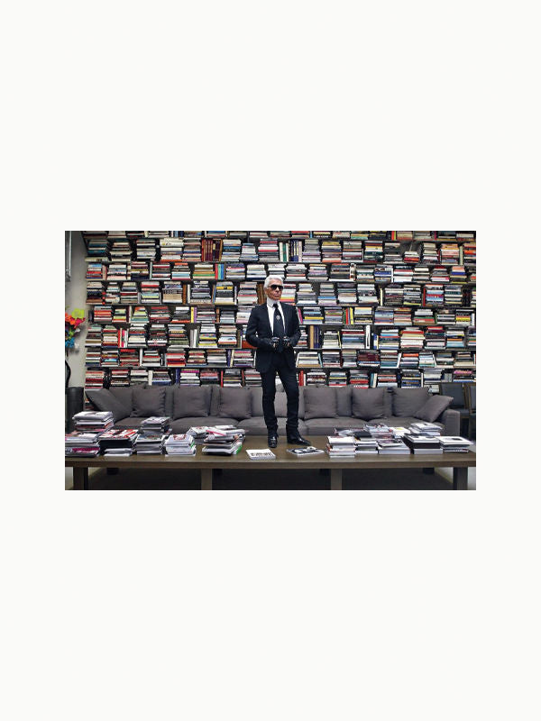 A person dressed in a black suit and sunglasses is standing on a couch in front of a large bookshelf filled with numerous colorful books, evoking the chic aesthetics of an art deco mansion. There is a table in front of the couch with stacks of books and magazines, including "Karl Lagerfeld: A Life in Houses" by Maison Plage, reminiscent of Karl Lagerfeld homes.