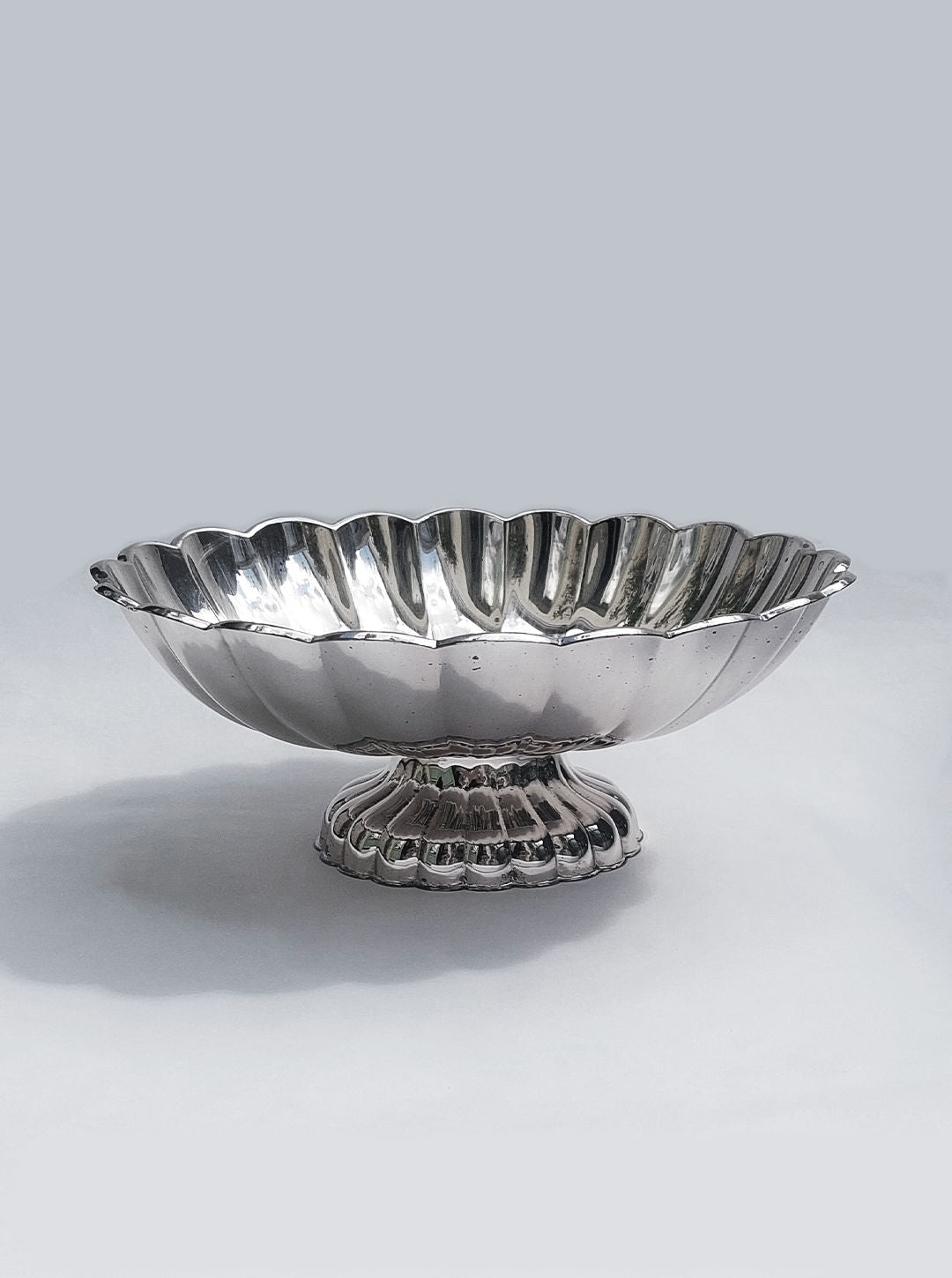 A silver Les Objoies Large Fruit Basket with a scalloped edge and fluted design is placed against a light grey background. The elegant piece features a wide, shallow basin supported by a short, ornate pedestal base, making it an exquisite centerpiece.