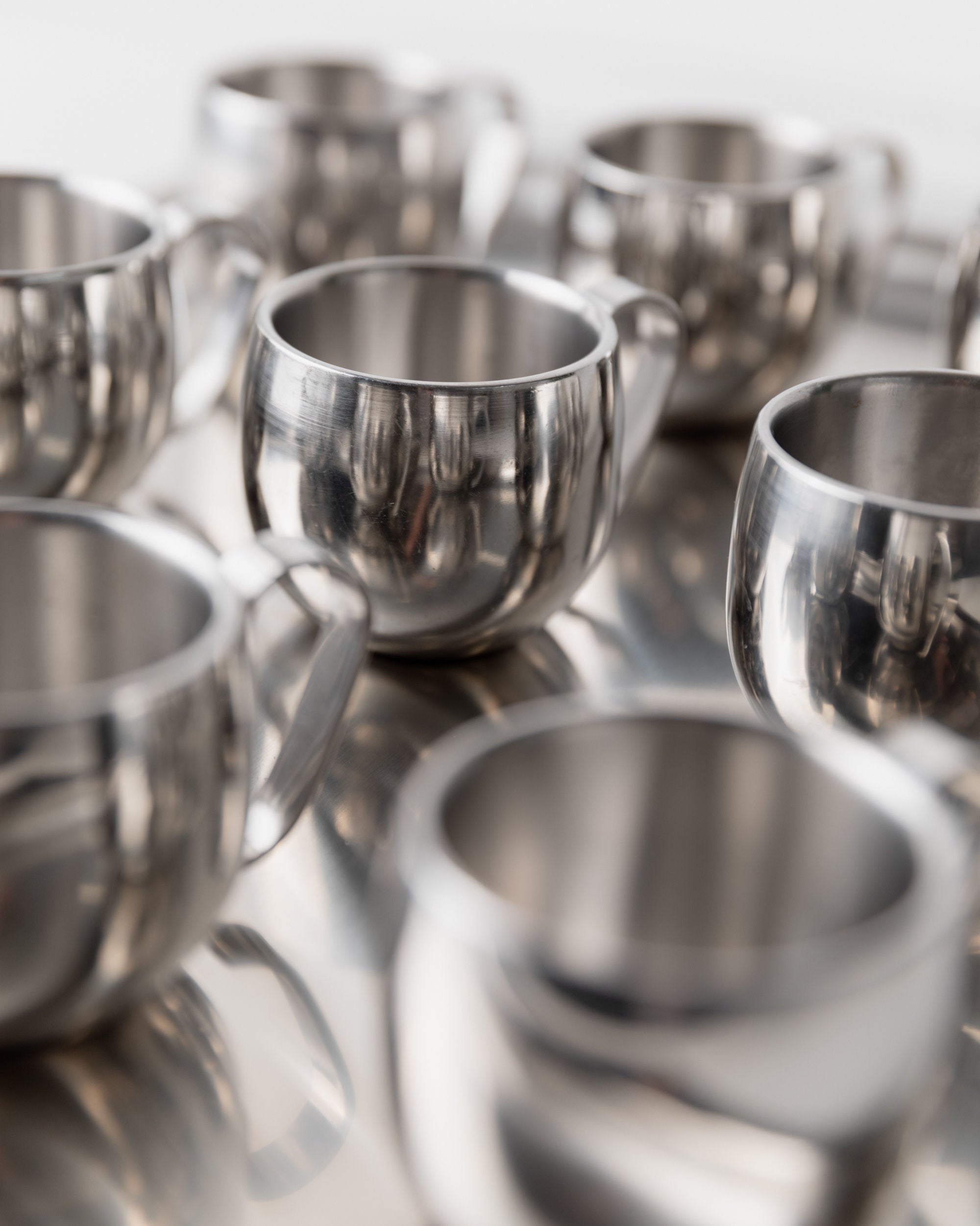 Close-up of several shiny stainless steel cups with handles from the Coffee set by IMCO 70s, by Bottega Jacobs. Their minimalist design creates soft highlights and subtle reflections against a blurred background.