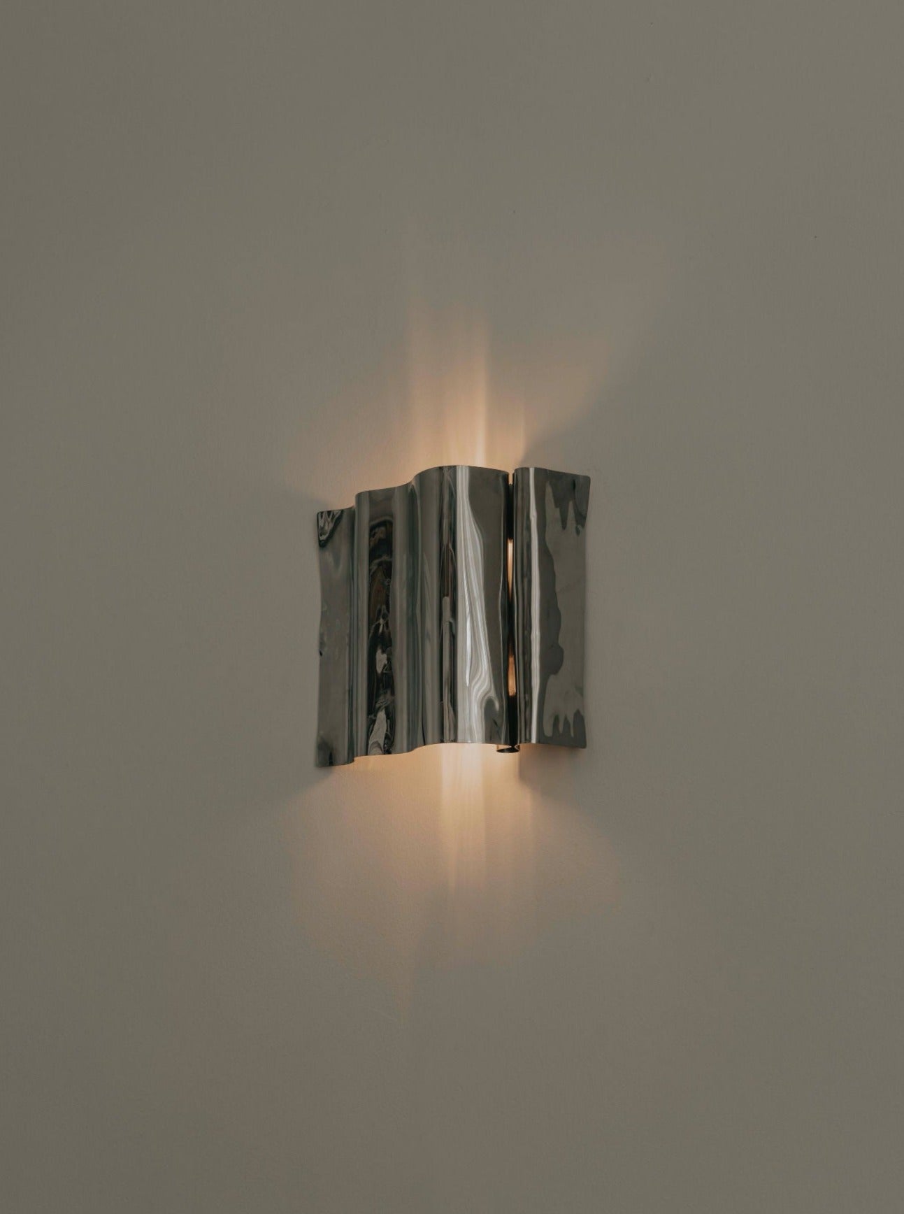 A contemporary wall sconce with a sleek, folding metal design emits a warm, soft light that creates subtle, patterned shadows on the beige wall. The handmade Psychedelic Wall Scone by Caia Leifsdotter boasts a shiny, reflective finish in polished stainless steel that enhances its unique organic shape and modern aesthetic.