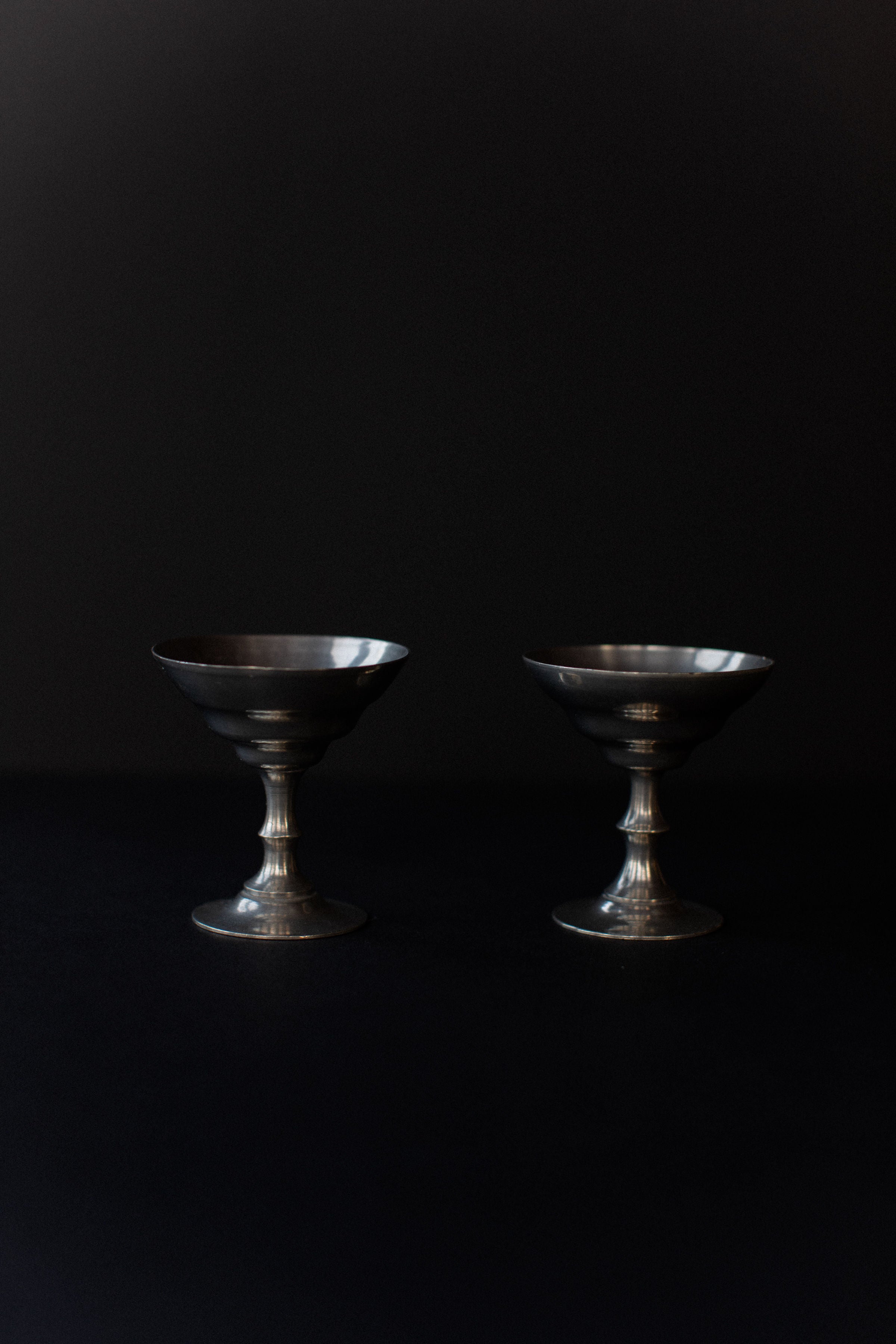 Displayed against a dark backdrop, two vintage-style Ice Cream Cups by Out For Lunch feature wide, shallow bowls and ornate stems. Their polished metal gleams under subtle lighting, with elegant lines reflecting Art Deco influence.