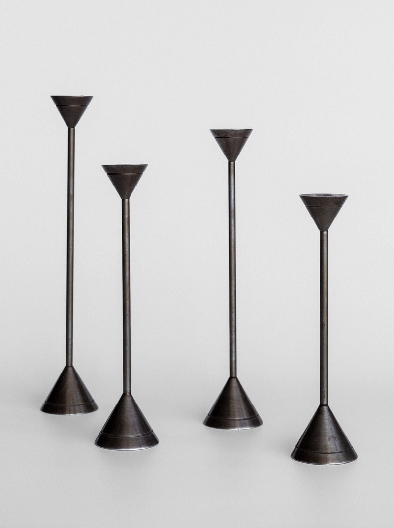 The Spigel Set of Four Postmodern Candle Holders are showcased against a light grey background. Each candle holder features a minimalist, solid metal construction with varying heights, conical bases, and slender stems leading to small, funnel-shaped tops designed to hold candles. Their sleek design exudes postmodern aesthetics.