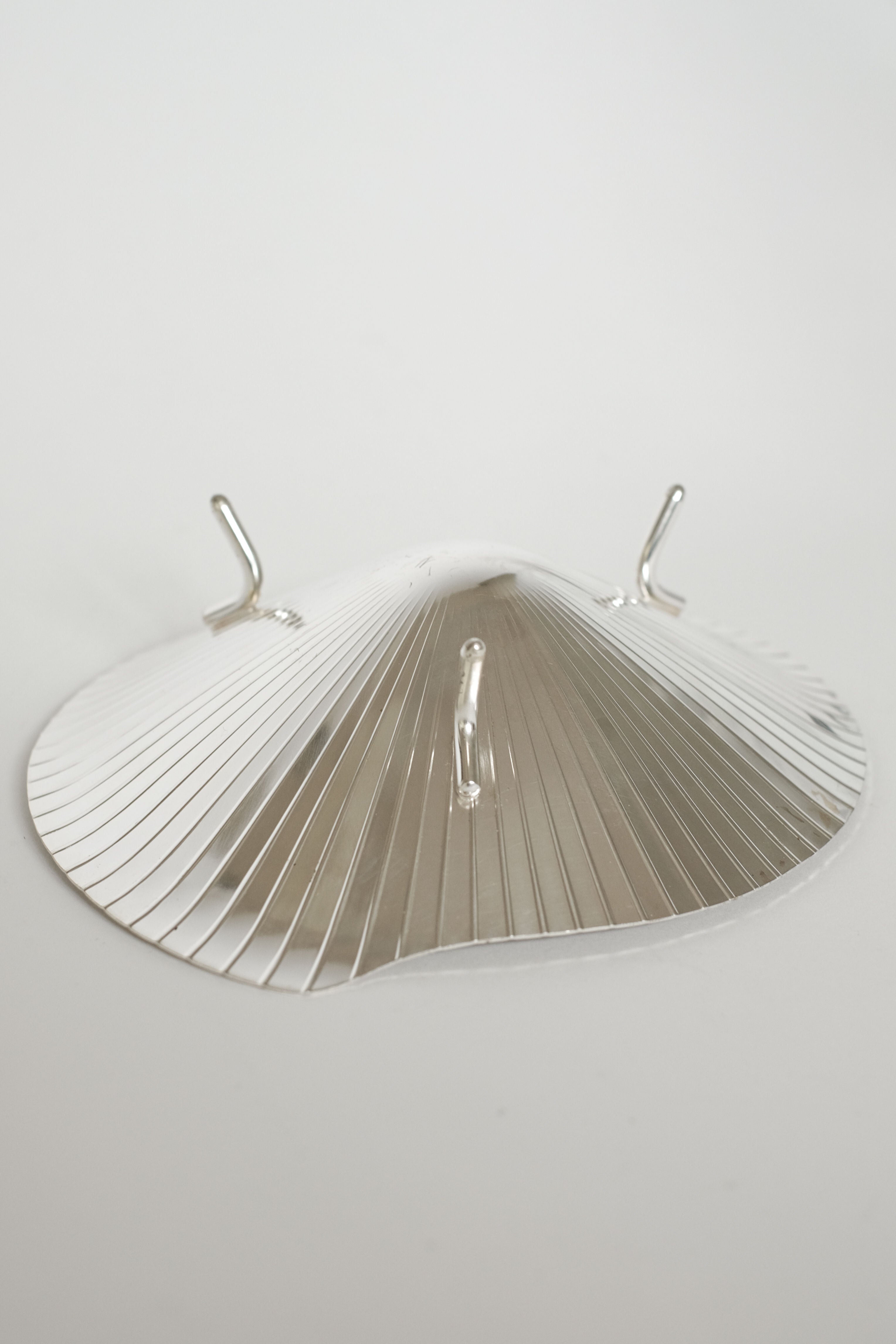The Silverplated Shell Dish WMF by septembre studios features a vintage metallic appeal with a ridged, lamp shade-like design. It elegantly rests on a plain white surface and includes three small hooks on top.