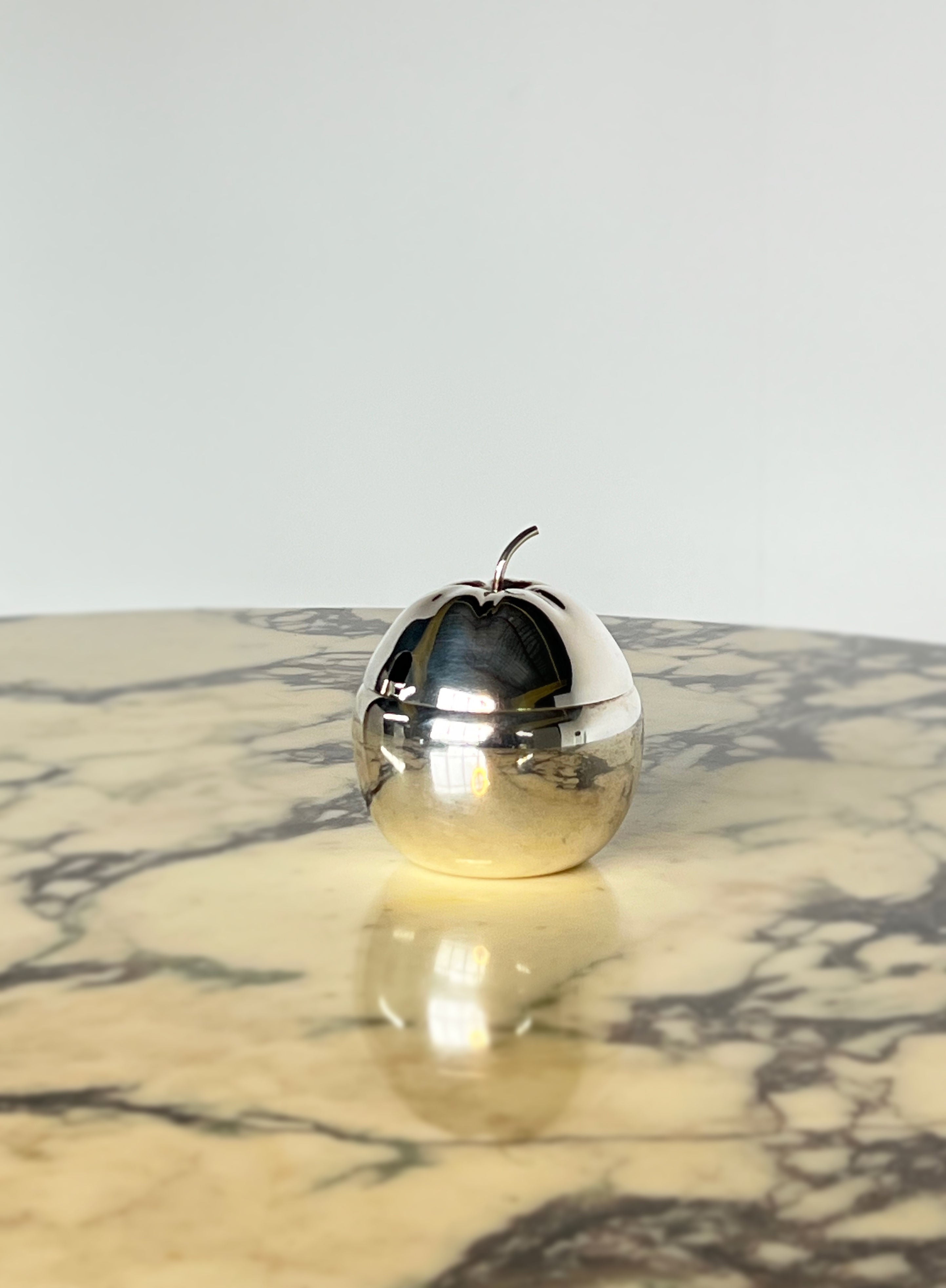 The Porter Studio Silver Plated Apple Jam Pot rests on a marbled surface, its polished exterior reflecting light and dark stone-like patterns. The softly blurred background enhances the apple-shaped container's reflective quality.