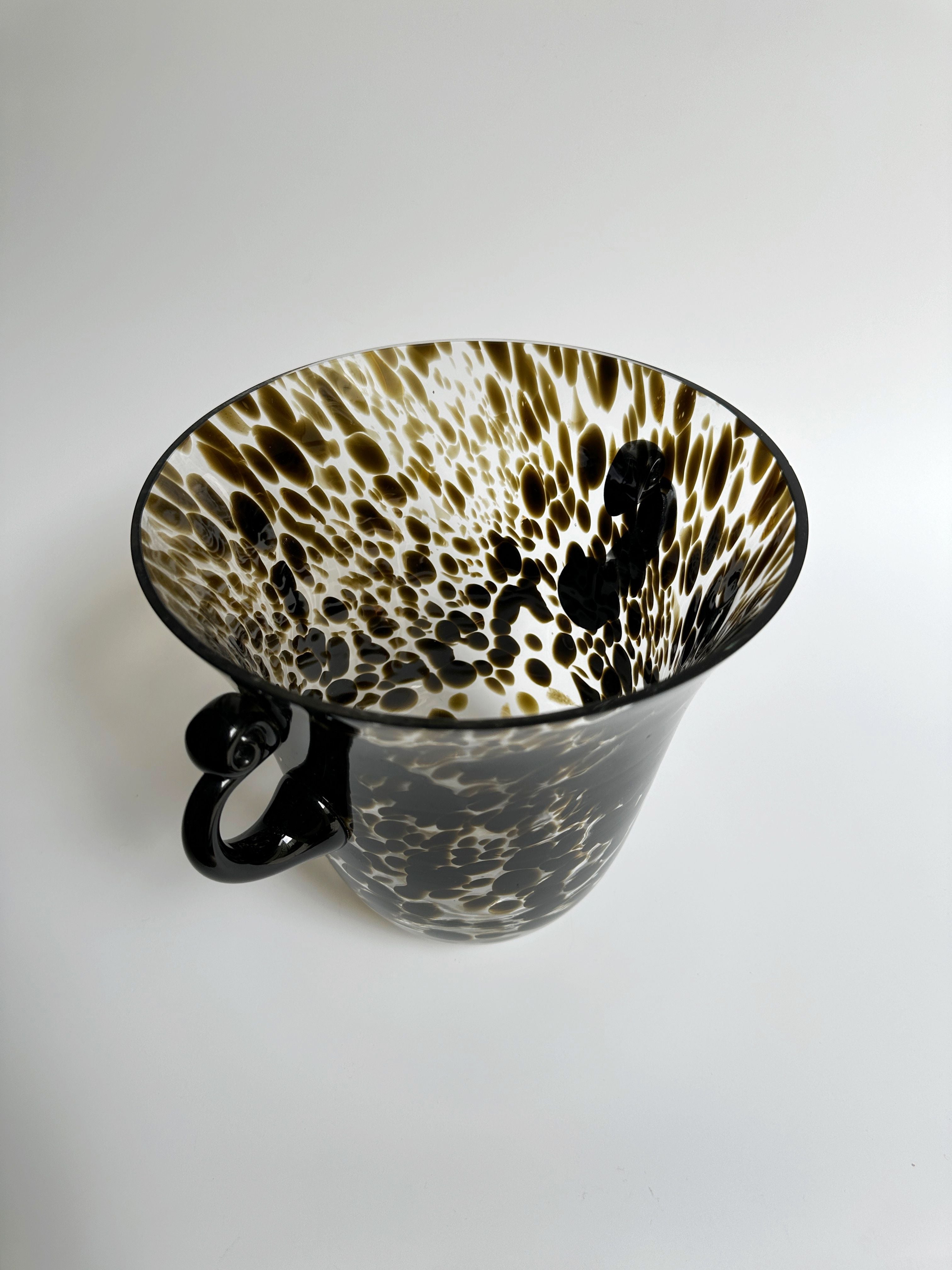 A striking glass cup designed with an abstract black and white spotted pattern, similar to the Dodo Vintage Art Deco Champagne Bucket/Vase Tortoise Glass. The interior design mimics ink drops spreading outward, creating a captivating artistic effect. This beautiful cup features a delicate, clear handle and rests on a plain white surface.