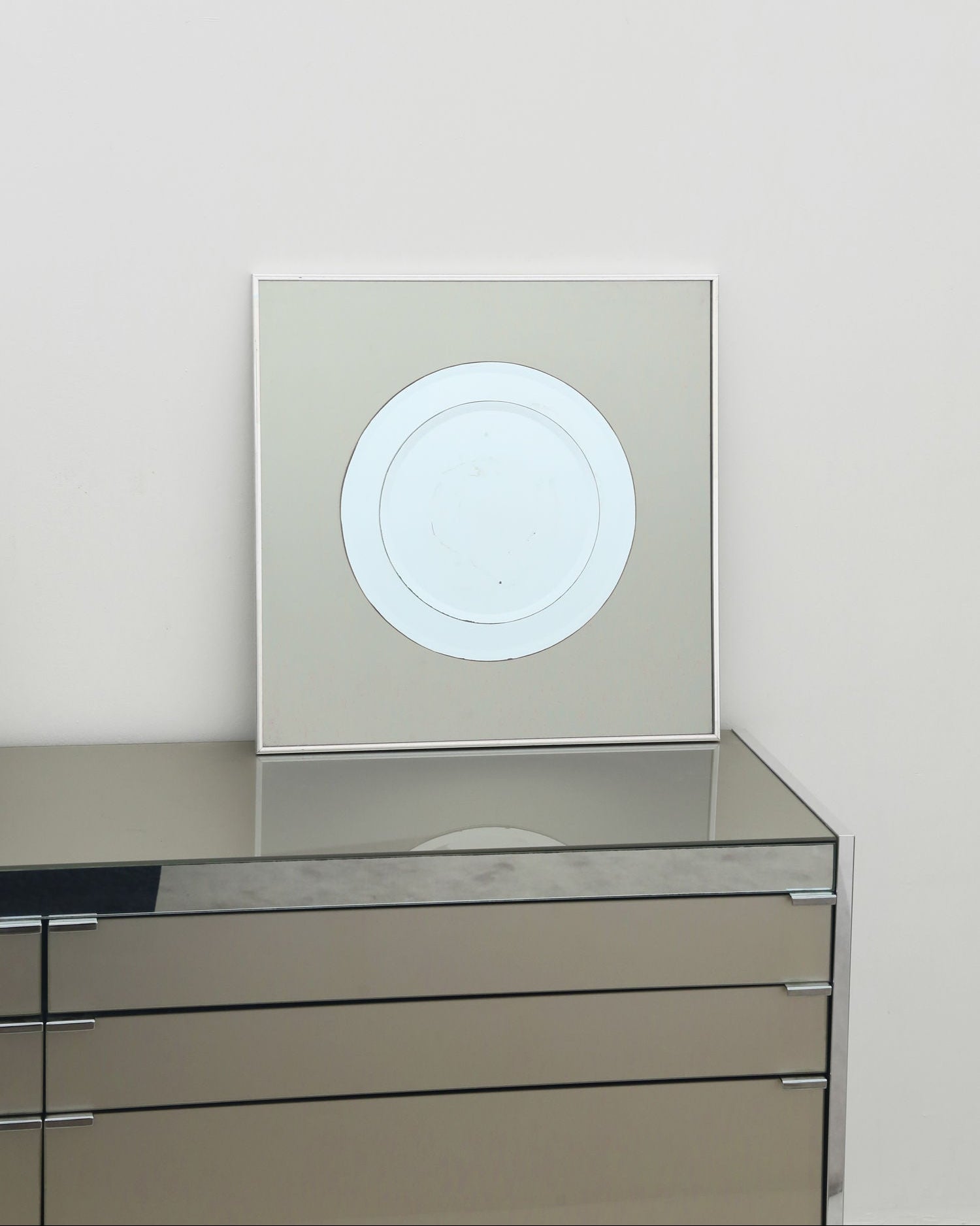 A minimalist circular artwork sits atop Villa's 1970’s Mirror dresser, a sleek two-tone piece with three drawers, set against a plain white wall.