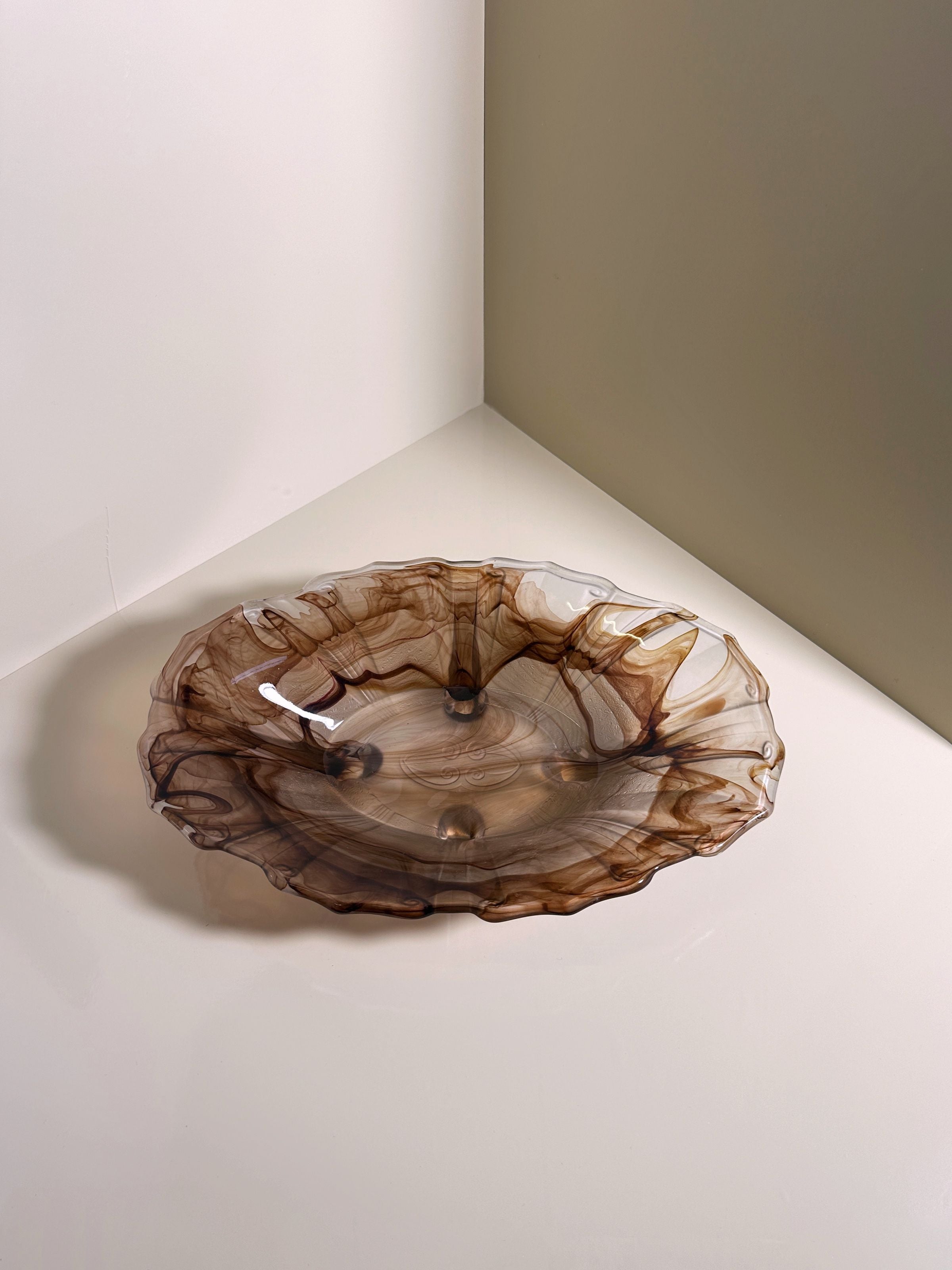 A translucent, wavy glass bowl with a smoky brown tint from Dodo Vintage's German Art Deco Cloud Glass collection circa 1930 rests on a reflective surface in a minimalist setting, its delicate edges casting an artistic shadow.