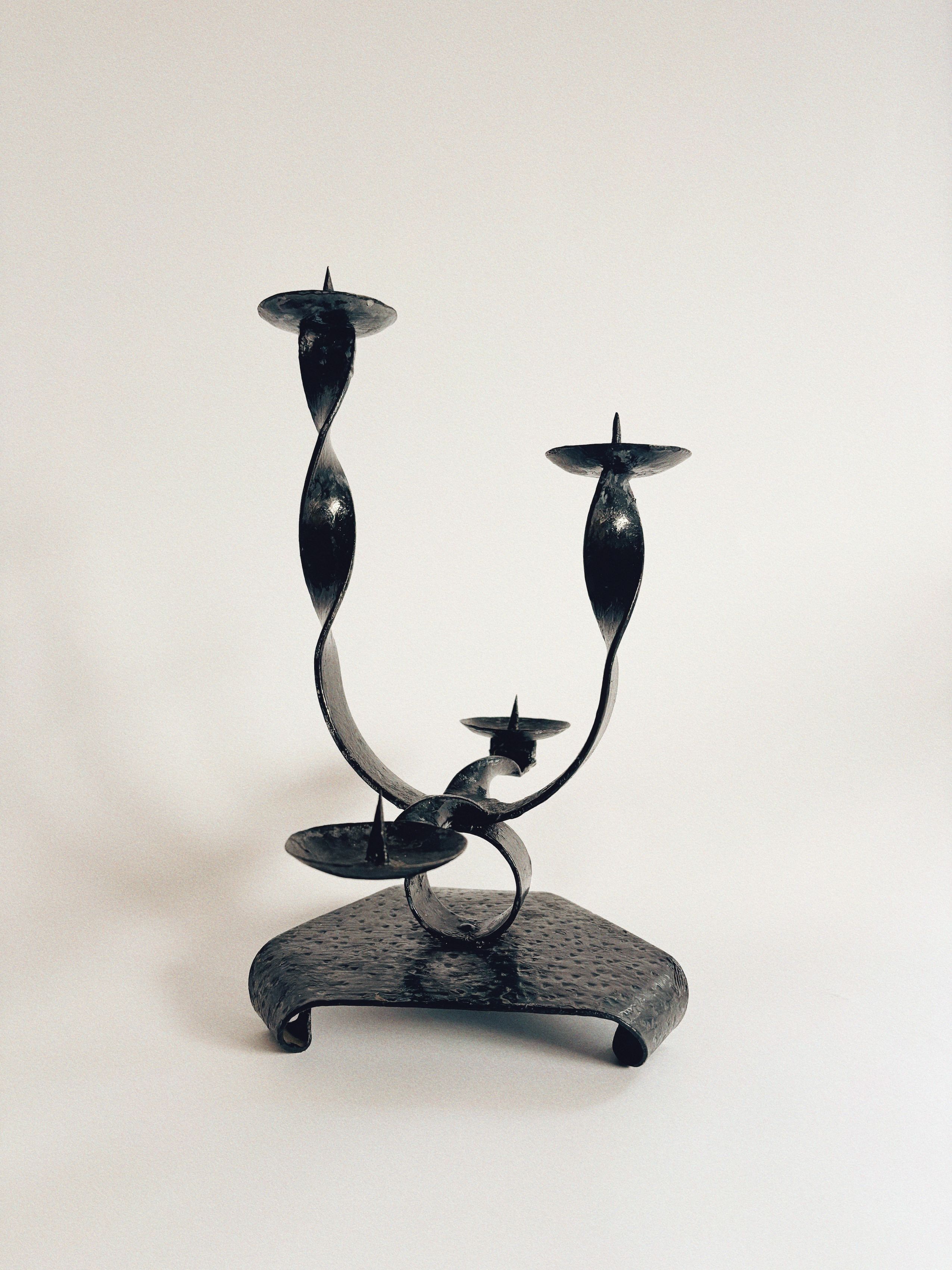 The Huerto Black Metal Candlestick showcases vintage artistry with four twisted ribbon-style arms and a sturdy, hammered base. Each arm has a flat circular surface for candles, all elegantly designed against a plain white background.