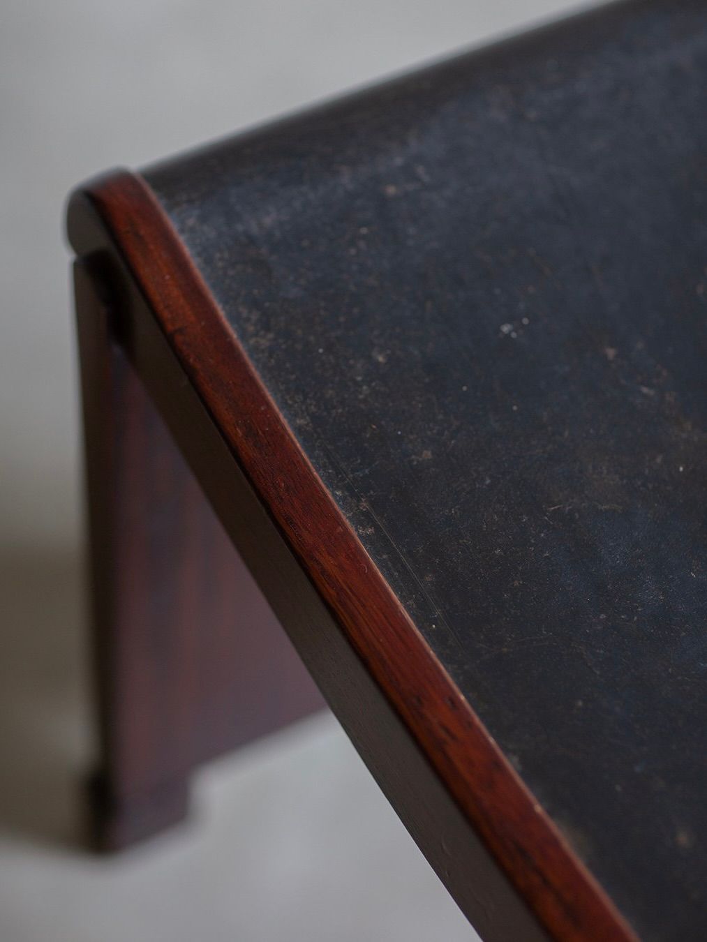 Close-up of a Pierre Chareau - Curule / Model No. MT 1015 Stool by Bicci de' Medici, featuring a vintage wooden corner with a dark, polished top and rich brown edges in Art Deco style. The worn surface adds character, set against a softly blurred background.