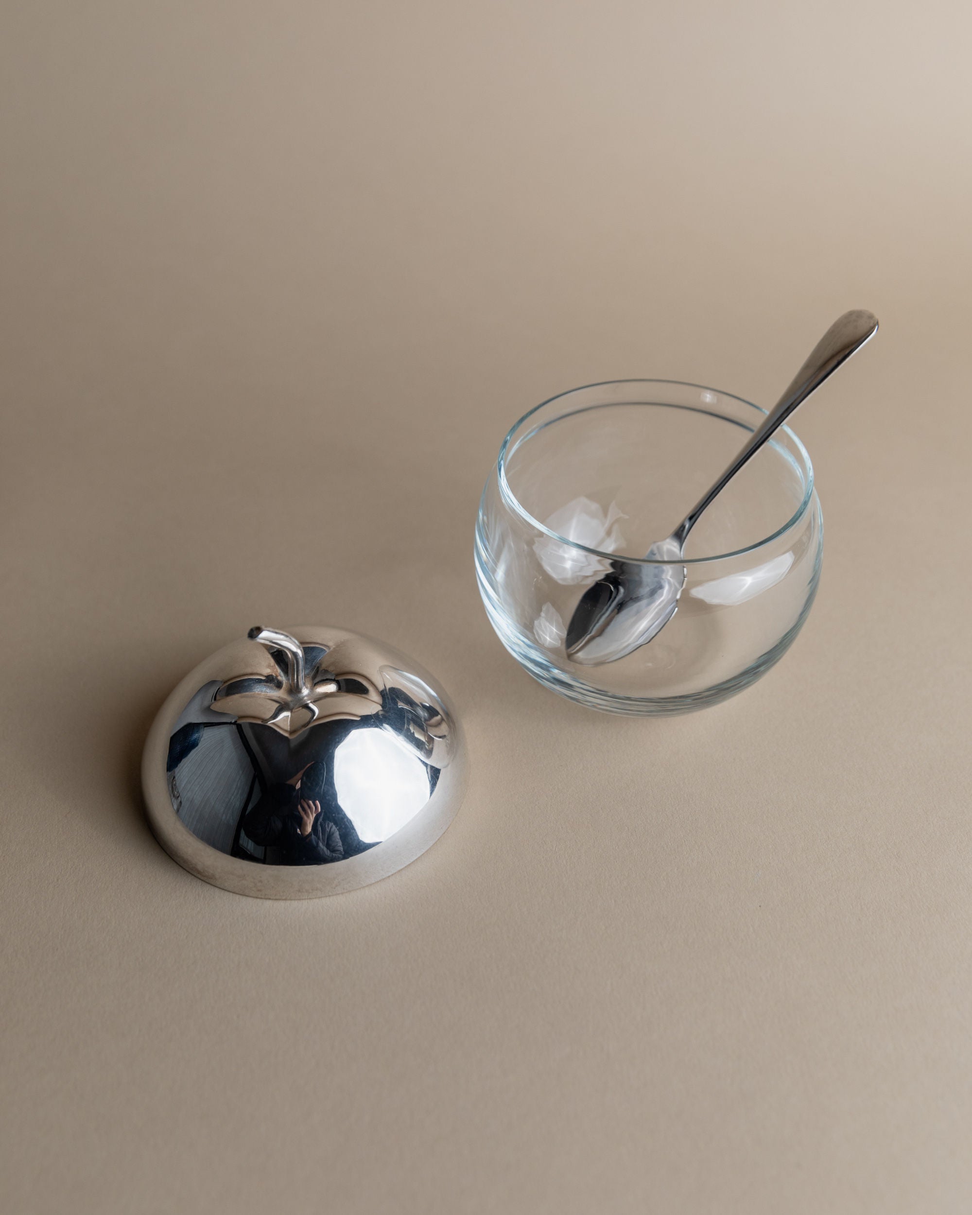 The Bottega Jacobs Silver & Glass Apple Jar, featuring a glass bowl with an internal spoon, sits on a beige surface and is paired with a silver-plated lid, complete with a handle shaped like two connected spheres.