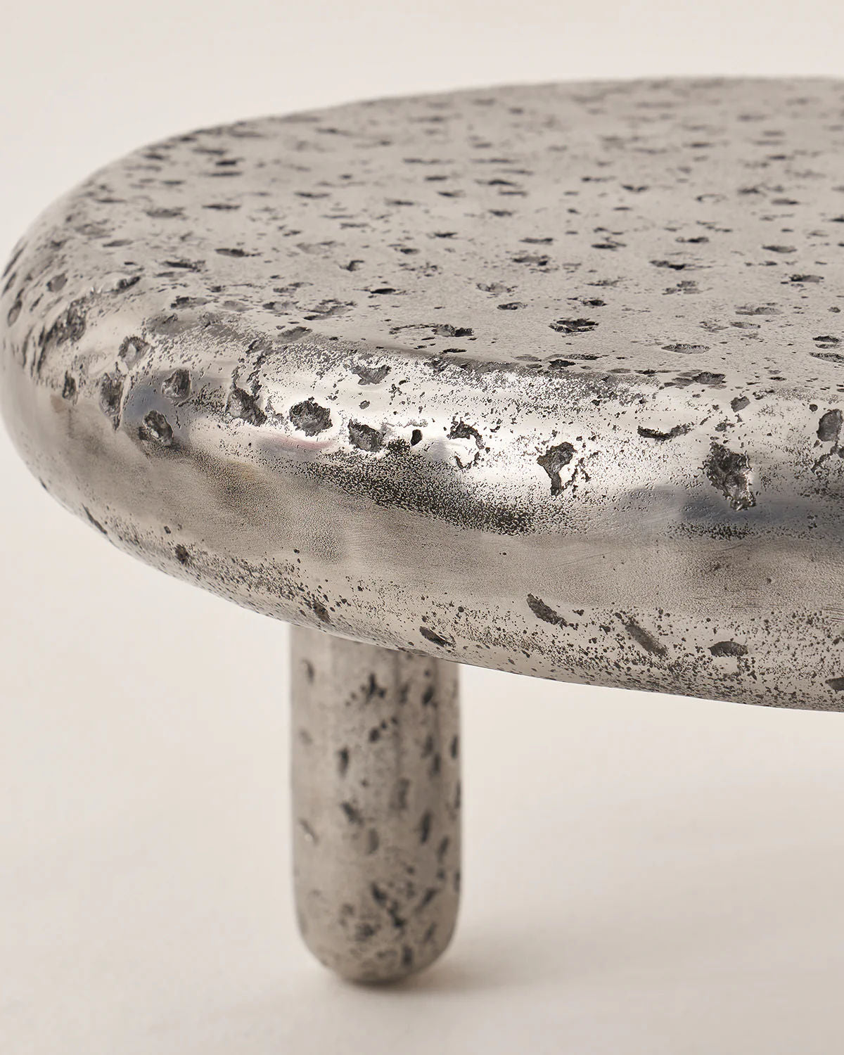 The Crater - Set of 2 by LMNOH features a close-up of a round, textured metallic stool top speckled like a lunar surface, coupled with smooth cylindrical legs. Its distressed finish adds rustic charm, making it a modern, industrial celestial centerpiece for any room.