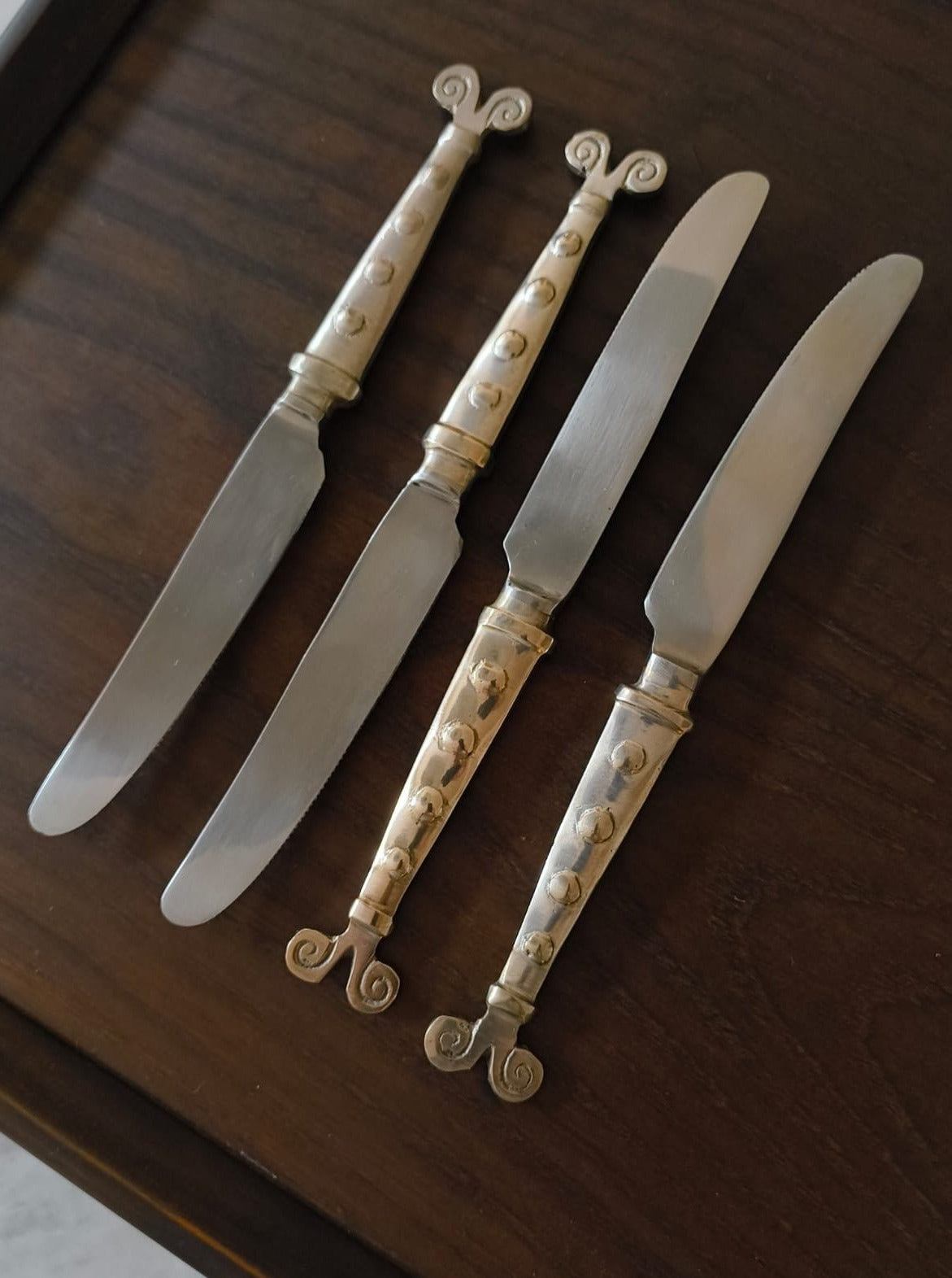 The Rare Vintage Cutlery Set by Médecine includes four artist-made butter knives, each with intricately designed handles featuring a spiral and dot pattern. Arranged neatly on a wooden surface, the shiny and smooth stainless steel blades offer a striking contrast to the elegant handle designs.