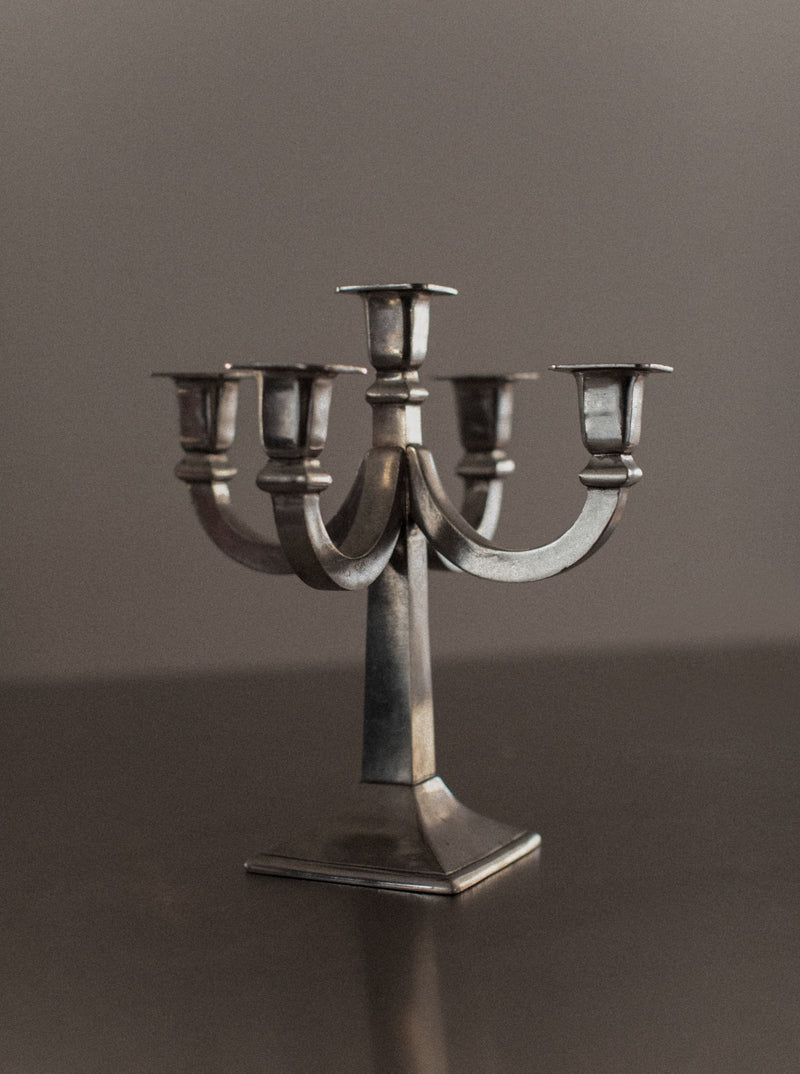 A vintage metal candelabrum with five candle holders arranged in a symmetrical design. Standing on a polished flat surface with a plain dark background, it boasts a slightly tarnished appearance, adding to its distinctive charm. This Mid-century Candleholder by Out For Lunch makes an elegant addition to any home decor.