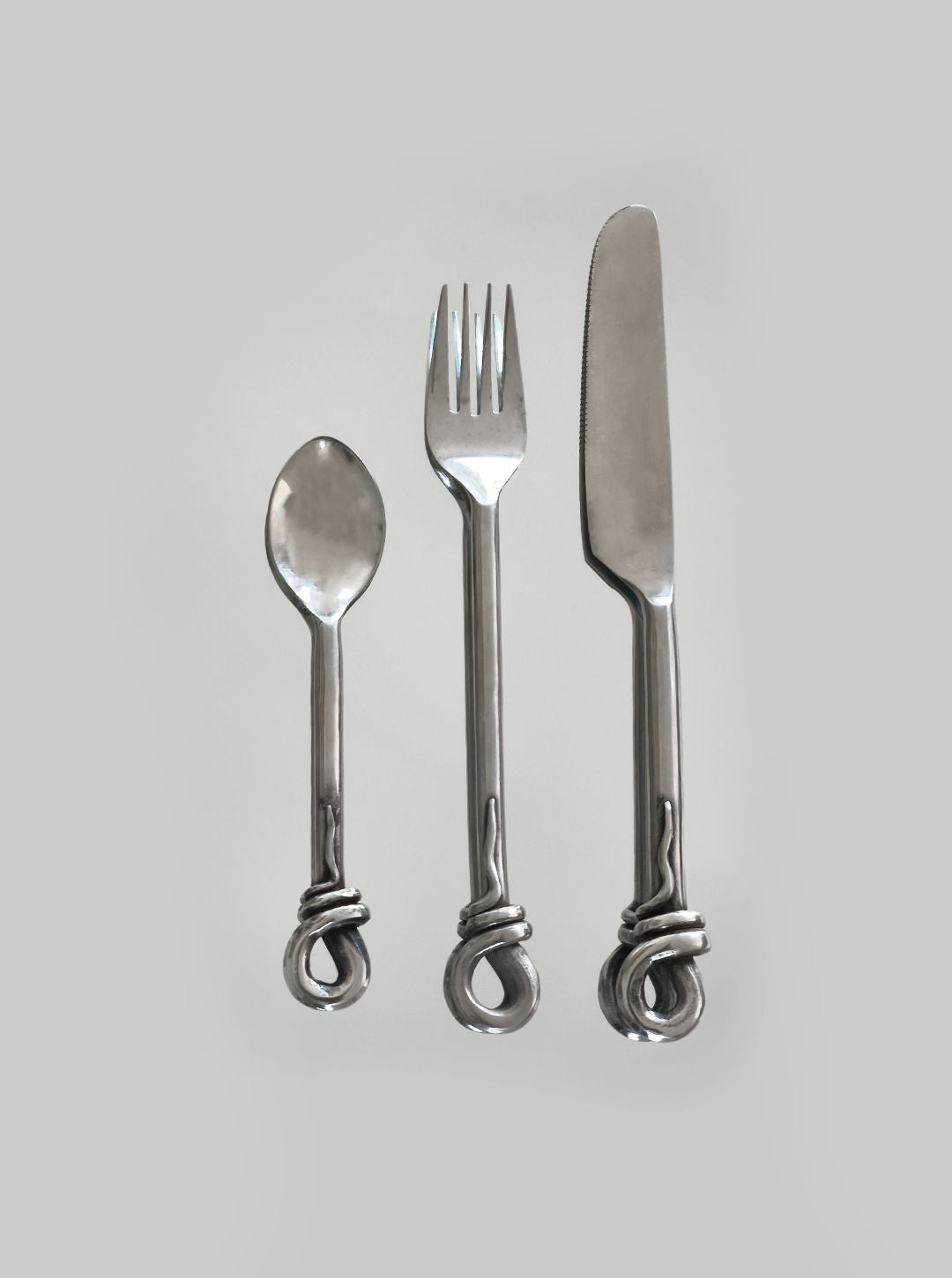 A captivating display on a gray background showcases the Set of 3 twist cutlery by Les Objoies, featuring a spoon, fork, and knife. Each piece is distinguished by its unique organic shape and elegant twisted looped handles.