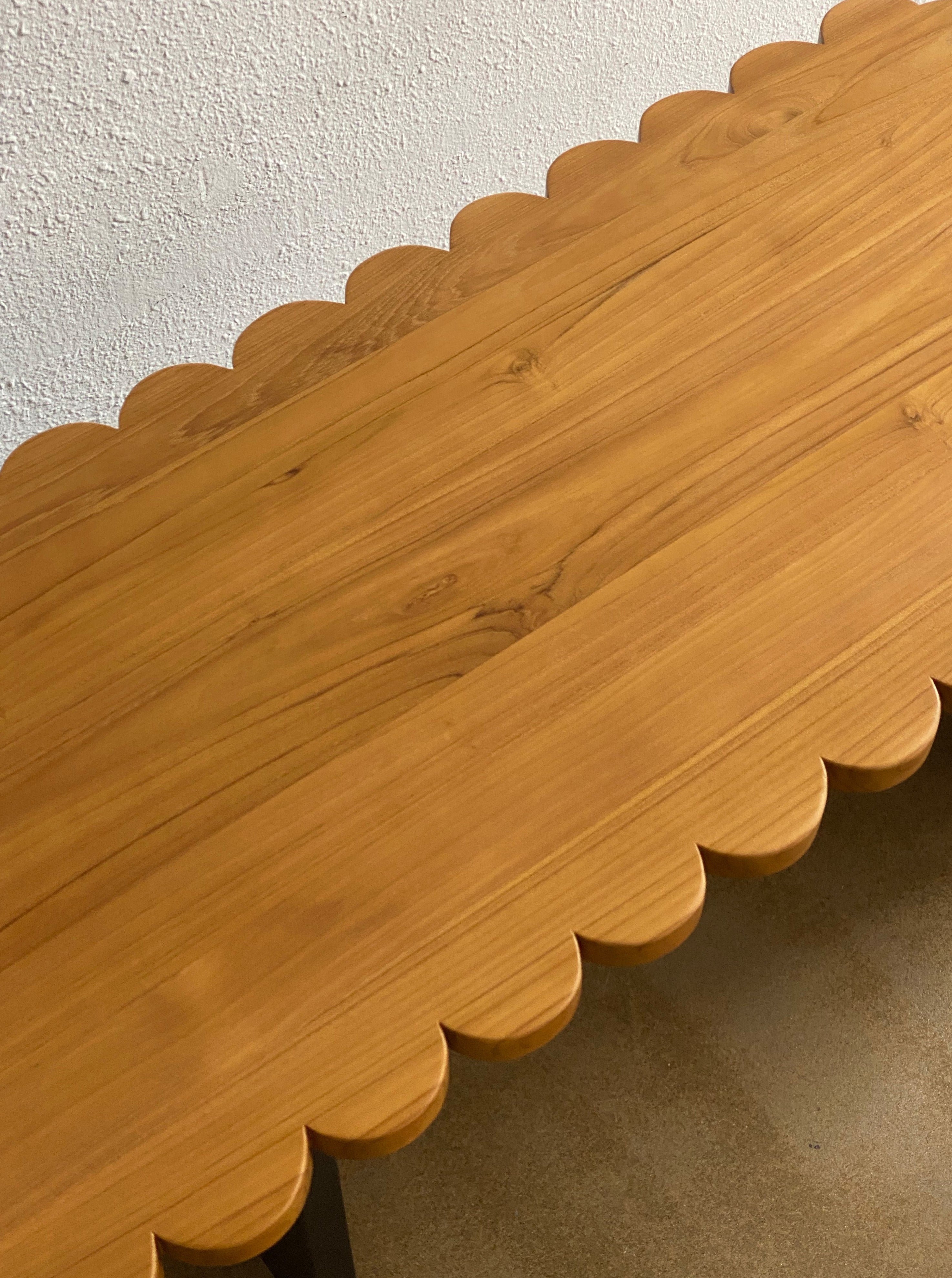 A handcrafted solid teak Biskut Coffee Table by Studio Kallang with scallop detailing is positioned against a textured white wall. The table’s surface displays a natural wood grain pattern, adding rustic charm to the piece. The flooring beneath the table is a light, neutral shade.