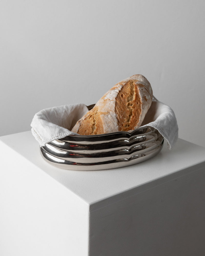 A loaf of crusty bread wrapped in a white cloth rests in the sleek, silver-plated "Bowl Silver-plated - Italy 1960s" from Bottega Jacobs. This elegant table piece sits on a white pedestal against a plain, light gray background.