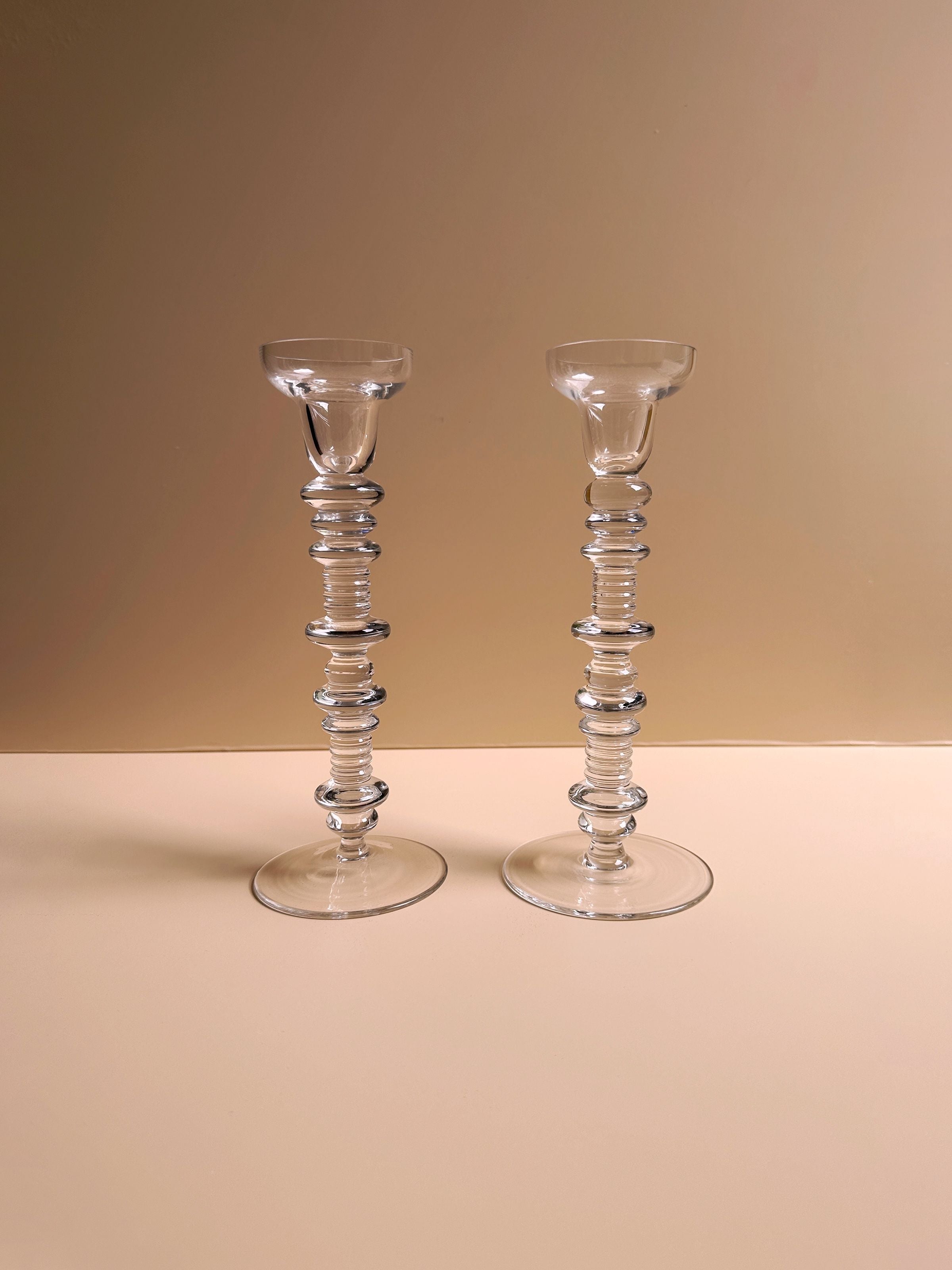 Two sophisticated glass candleholders, inspired by mid-century aesthetics, feature intricate stacked ring designs and stand side by side against a beige background. These clear, reflective pieces are part of the Set of 2 Glass Candleholders by Peill & Putzler from Dodo Vintage.