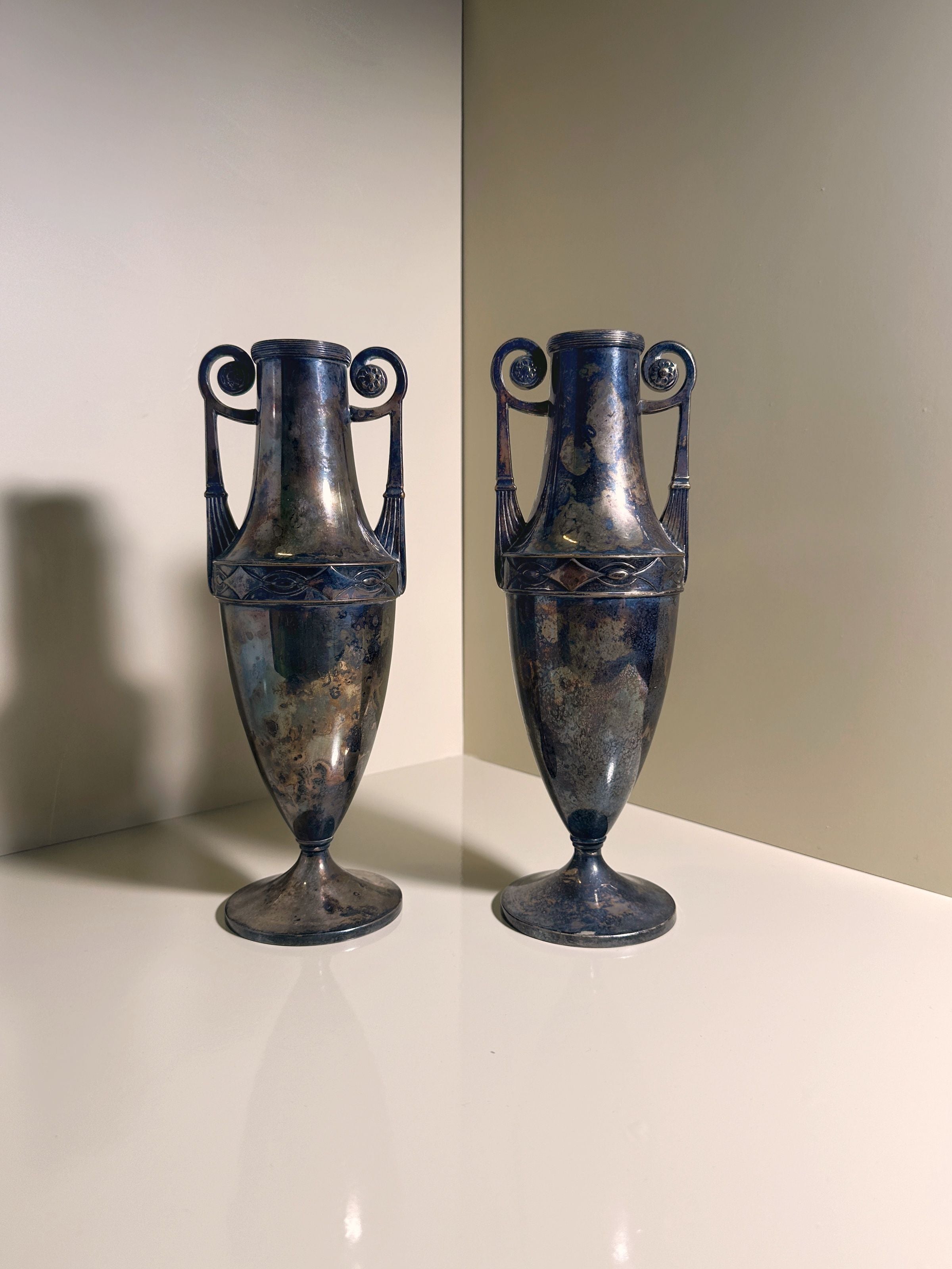 A pair of Dodo Vintage's silver-plated Art Nouveau Amphora from 1890 stand side by side on a reflective surface. Featuring intricate handles and classic, slender forms, they are set against a light-colored wall with soft shadows, highlighting their antique elegance.