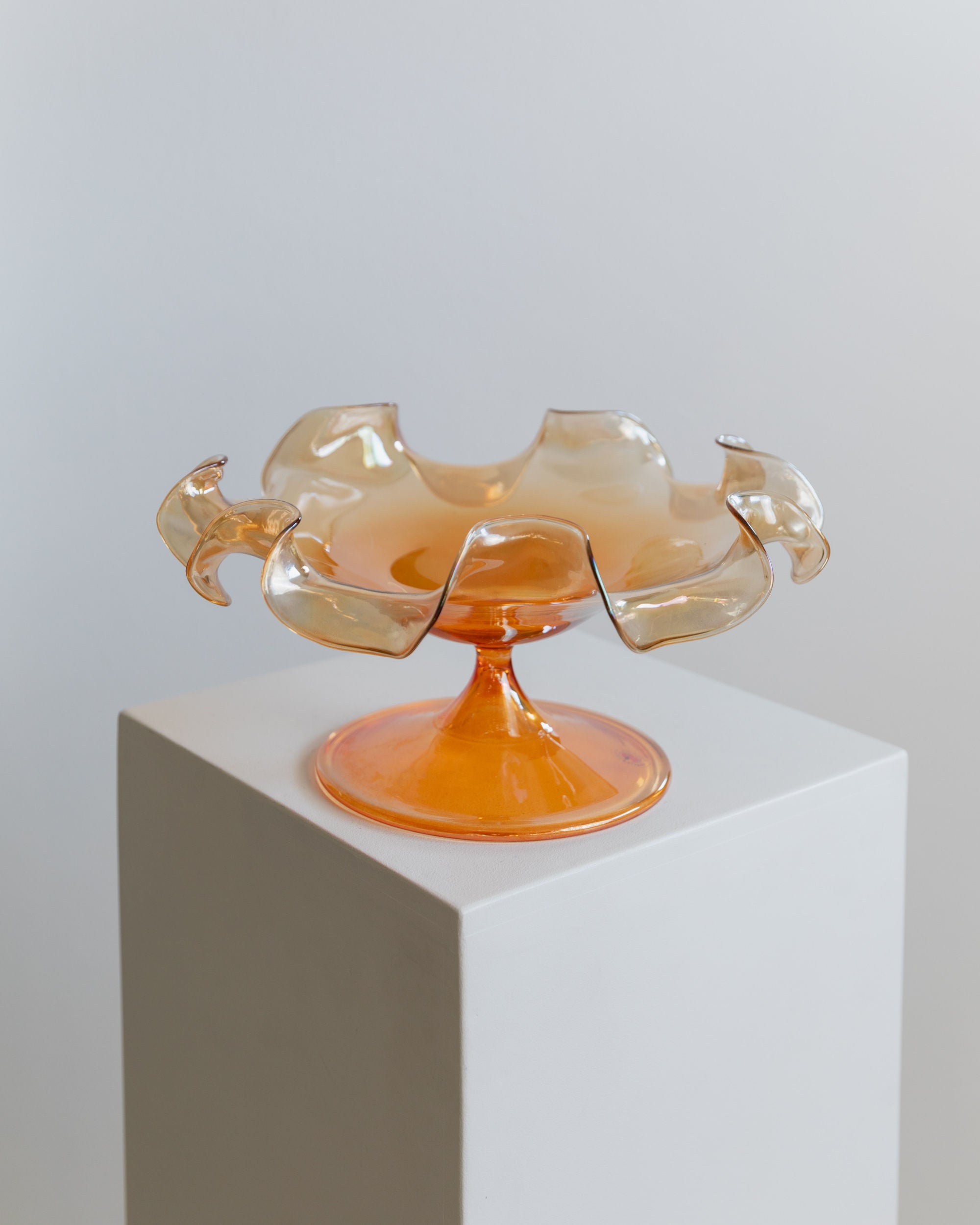 A decorative Murano Glass Bowl by Parise Vetro 70s from Bottega Jacobs, with its wavy edges, sits on a white pedestal. The translucent orange iridescence catches the light, highlighting its elegant design against a plain white wall.